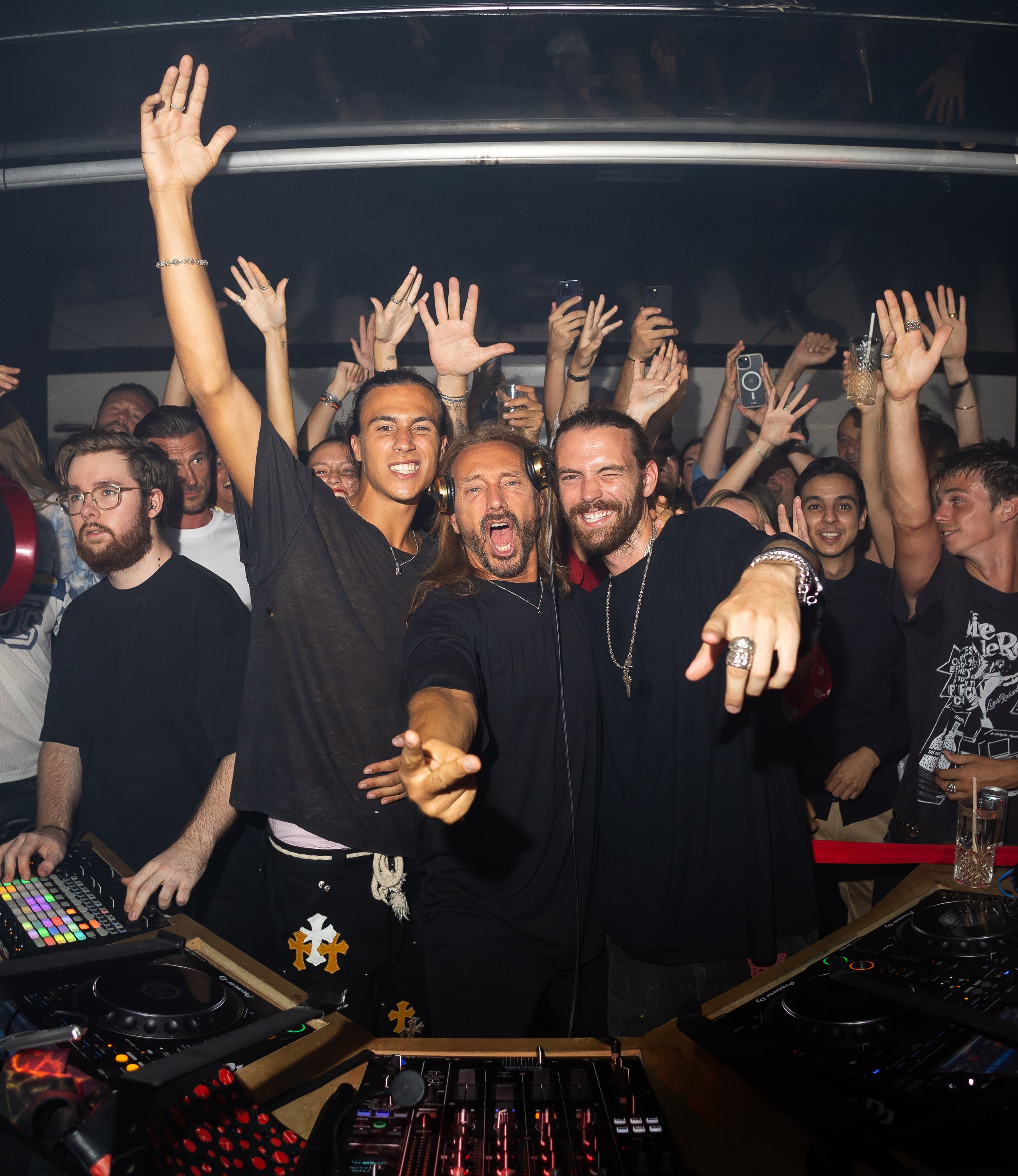 A group of people dancing at a party or club, with a DJ booth in the foreground, people raising their hands, some holding drinks, and a dark background.