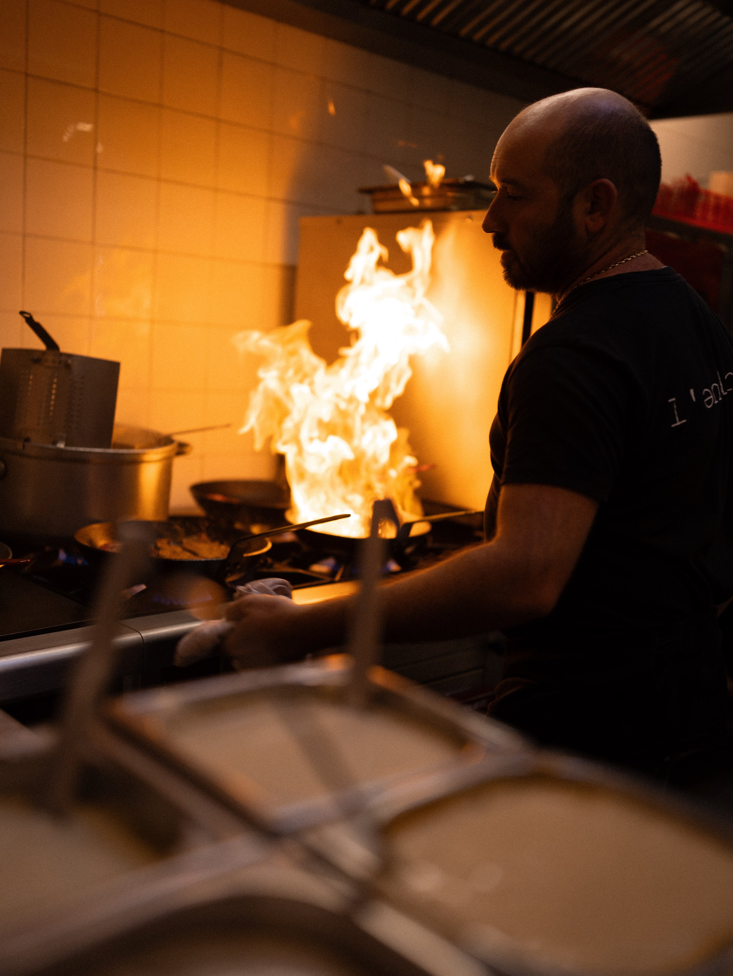 A man cooking in a kitchen with a large flame rising from a pan, with tiled wall and kitchen equipment visible.