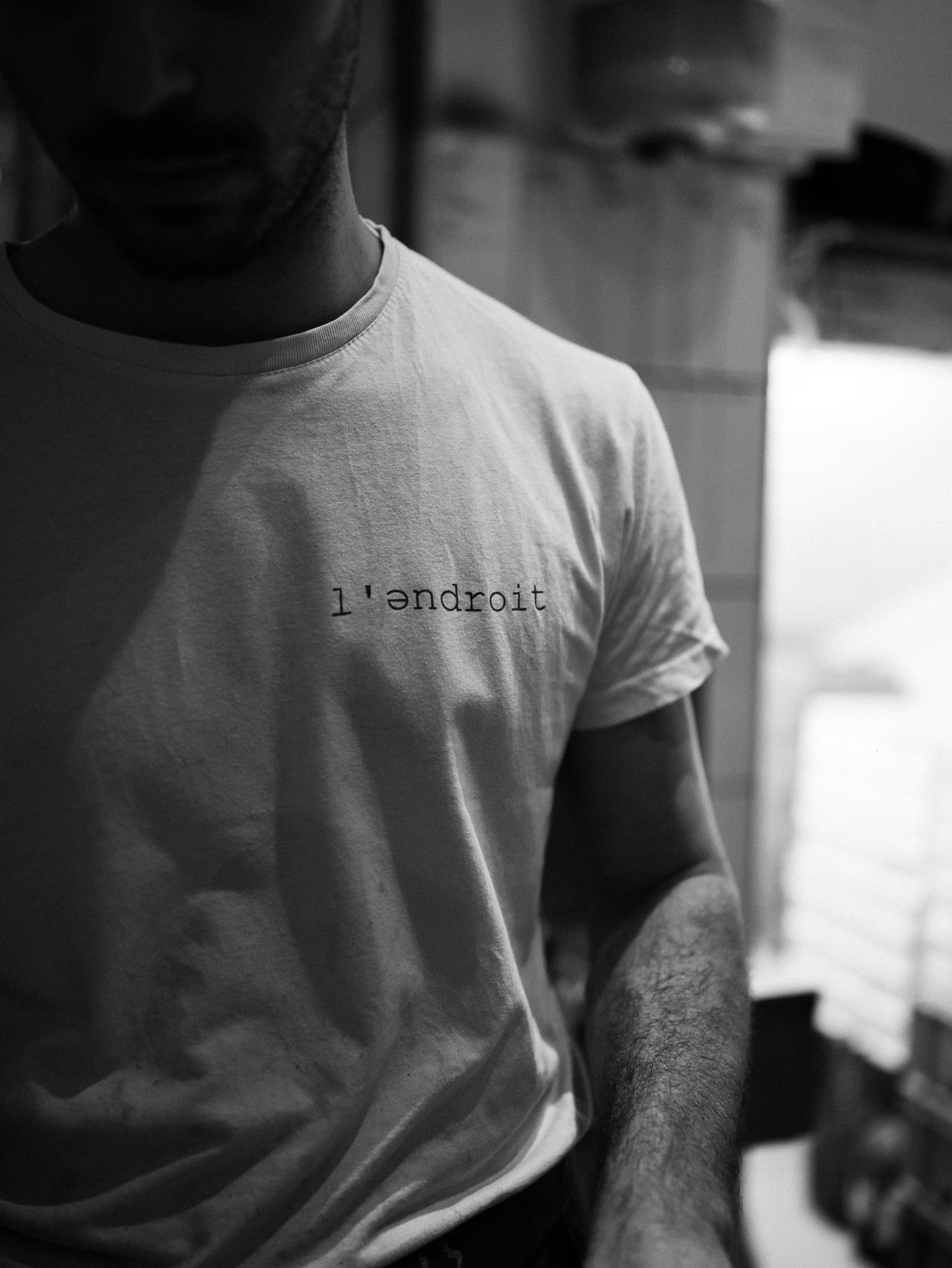 Close-up black and white photograph of a person's upper torso and part of their face, wearing a T-shirt with the text 'l'droid' printed on it. The person is looking downward, with a blurred indoor background.