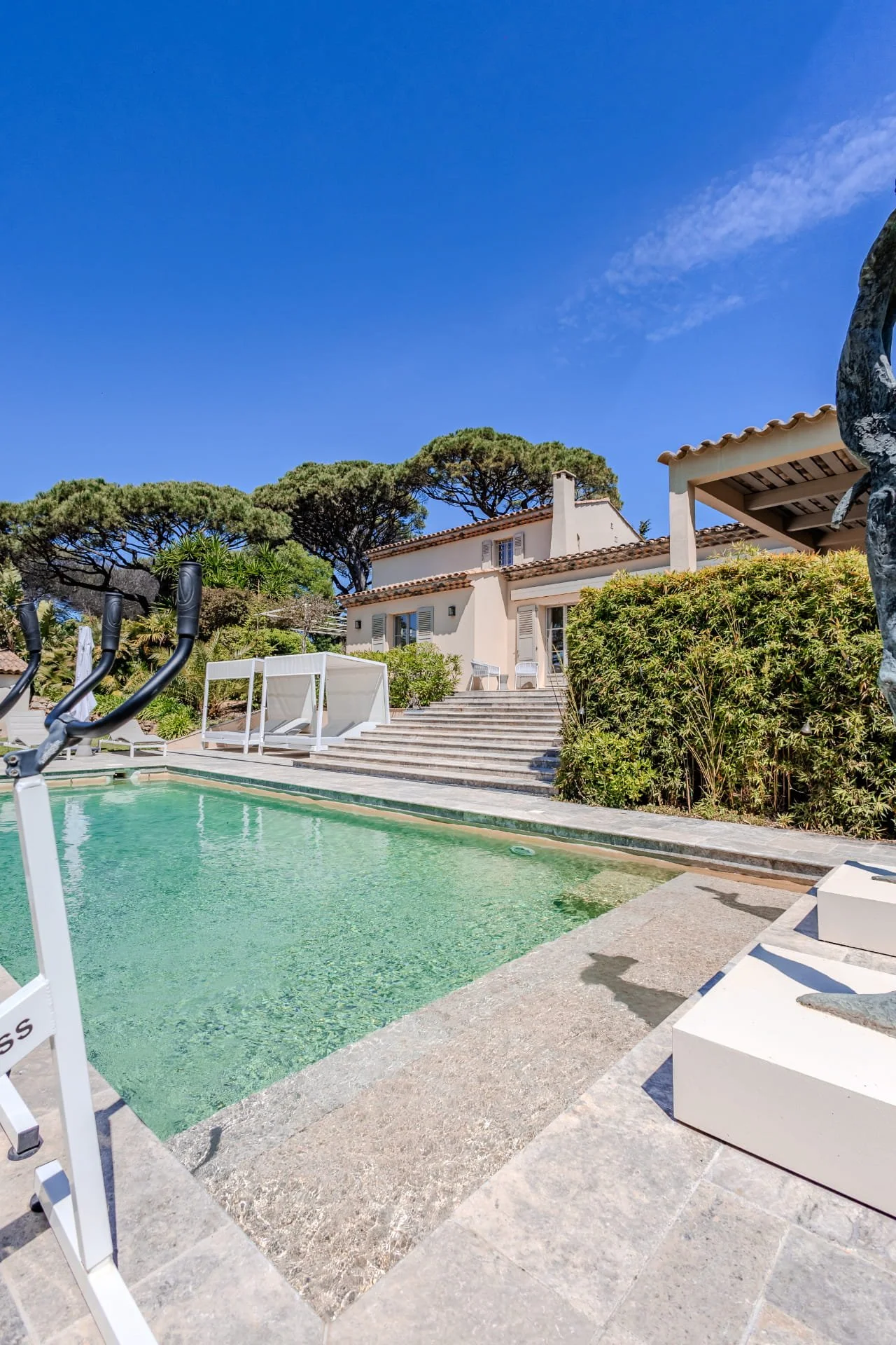 A backyard with a swimming pool, lounge chairs, and a white house with a tiled roof. There are trees and bushes around the house, and a clear blue sky above.