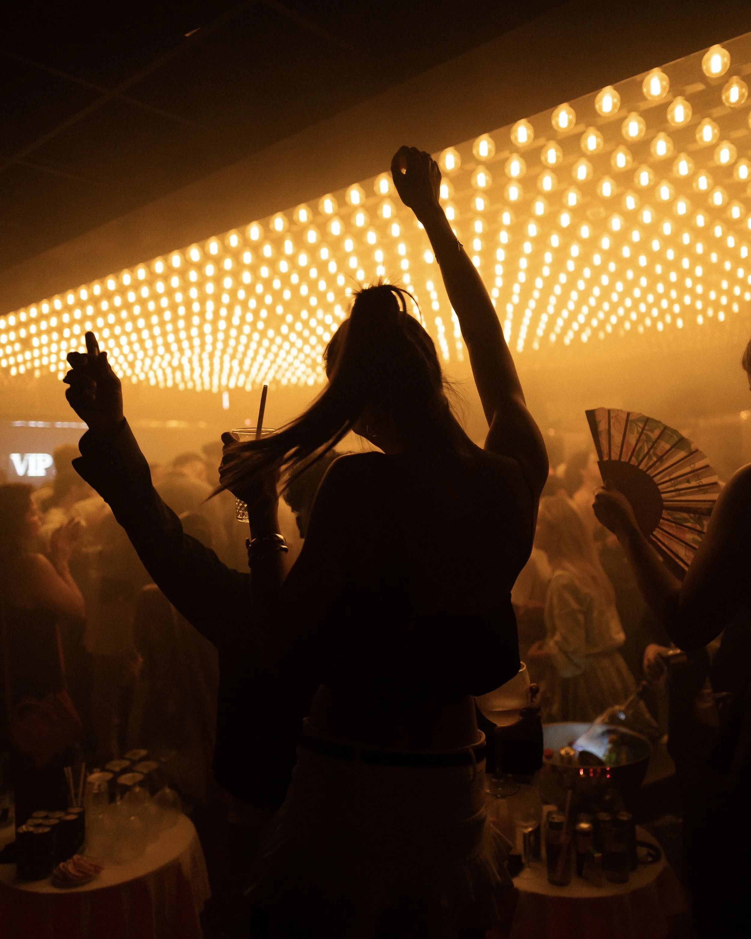 A woman with long hair is dancing at a party under warm yellow string lights, with other people and tables in the background.