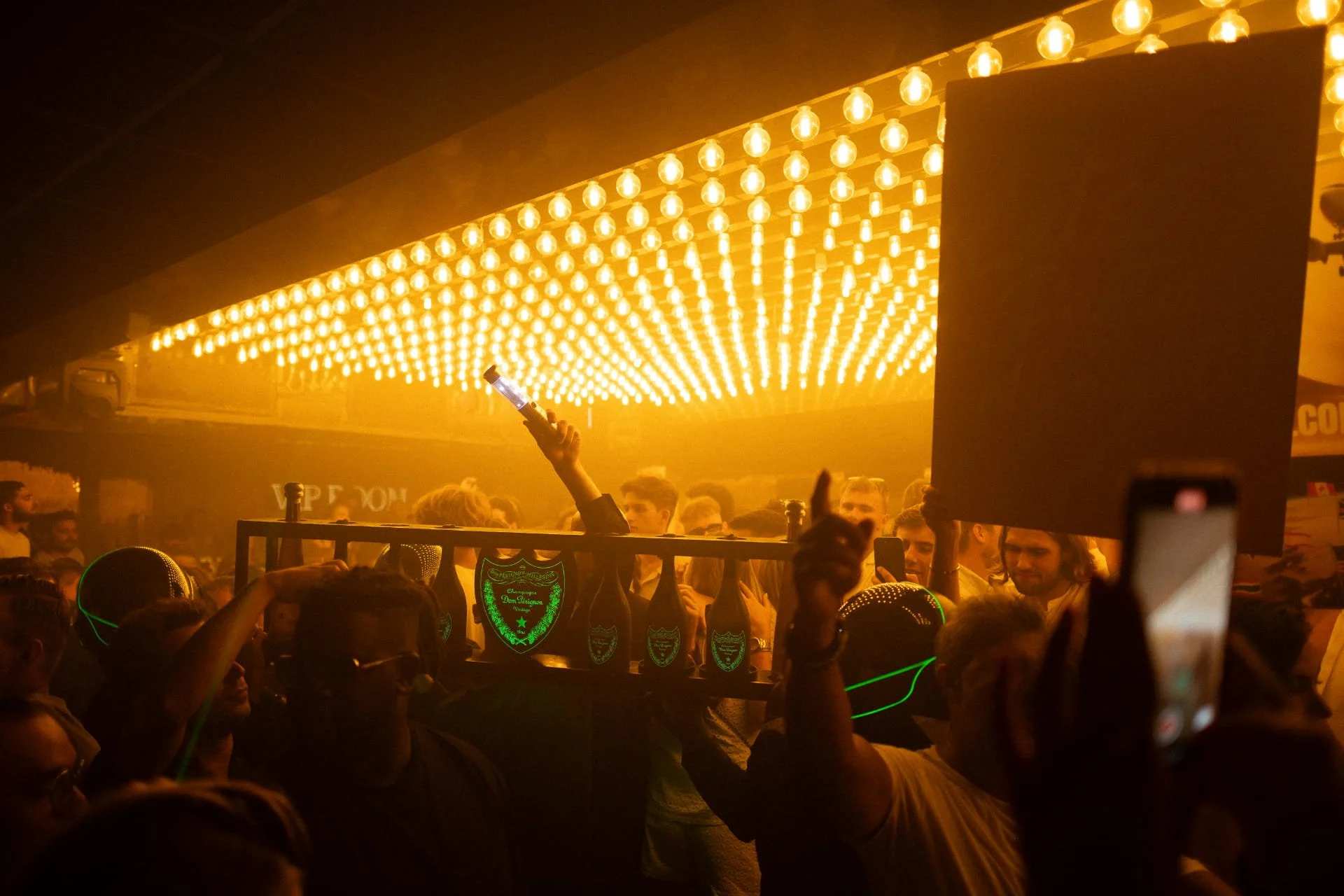 Crowd at a nightclub or concert with bright yellow hanging lights and people holding bottles and phones.