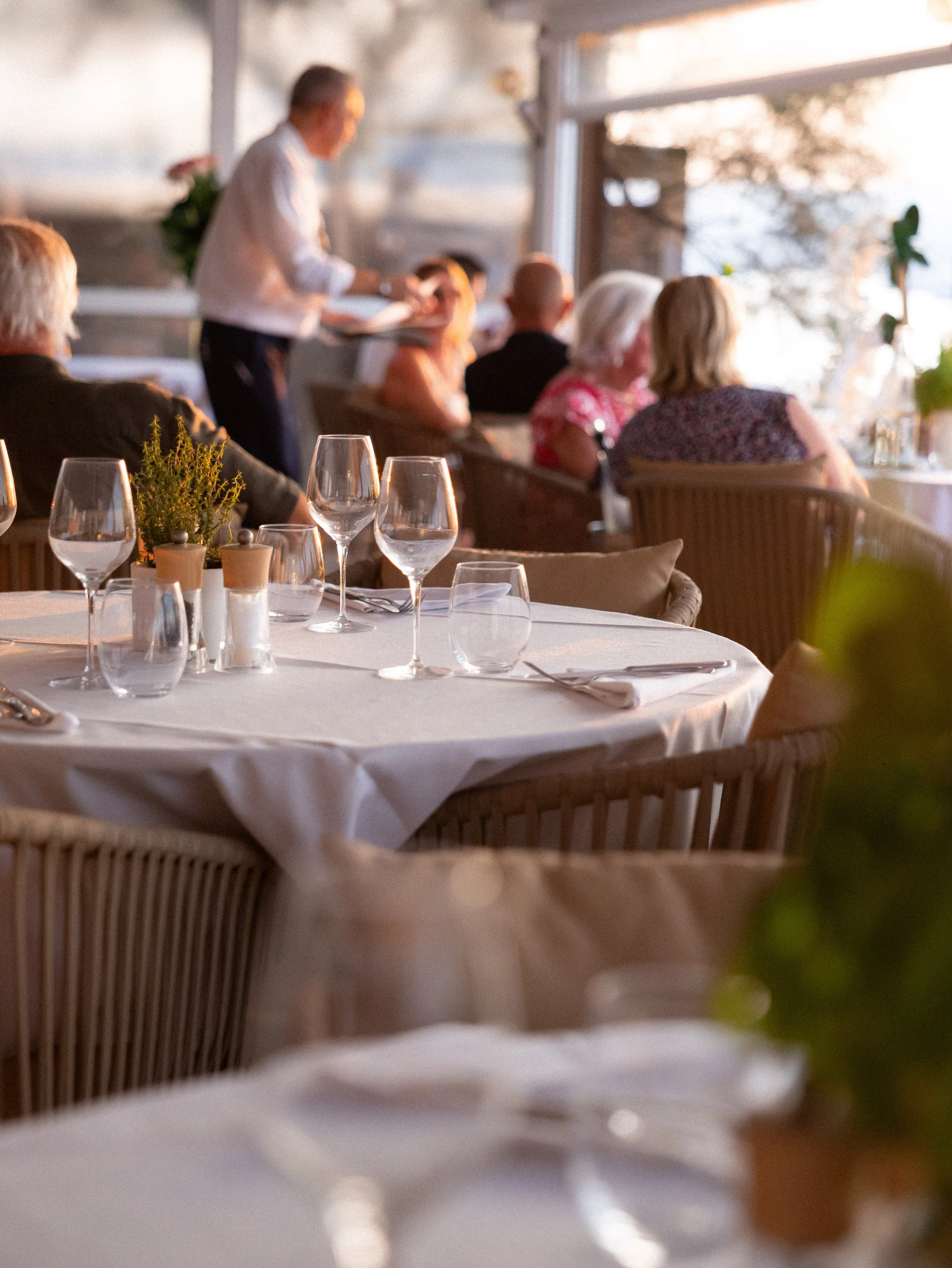 Blurred image of people dining at a restaurant with a view outside. The focus is on a table with empty wine glasses and a small plant in the foreground.