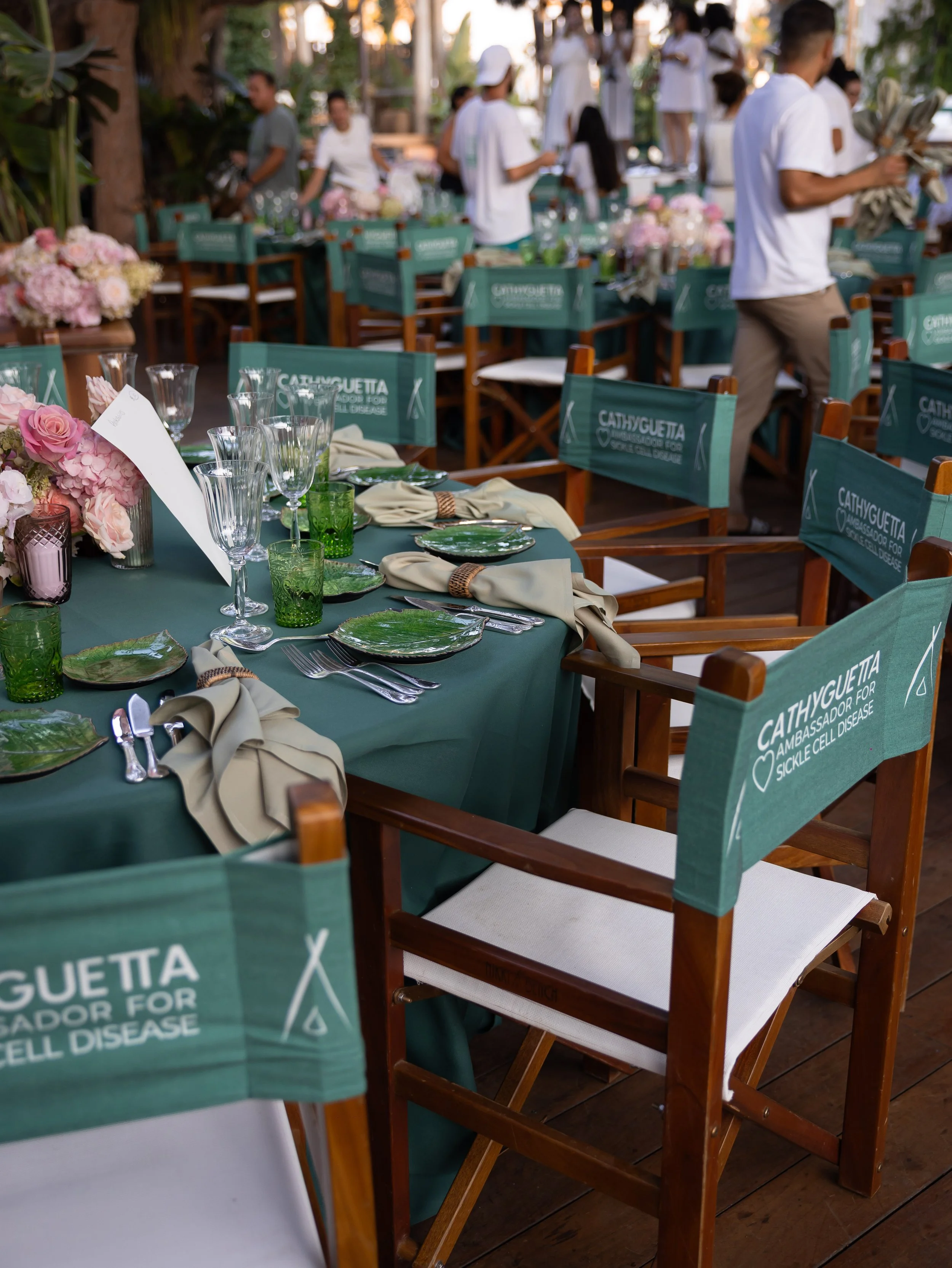 Wedding reception setup with tables decorated with flowers and glassware, surrounded by chairs with green covers that have white writing, in an outdoor setting with trees and people in the background.