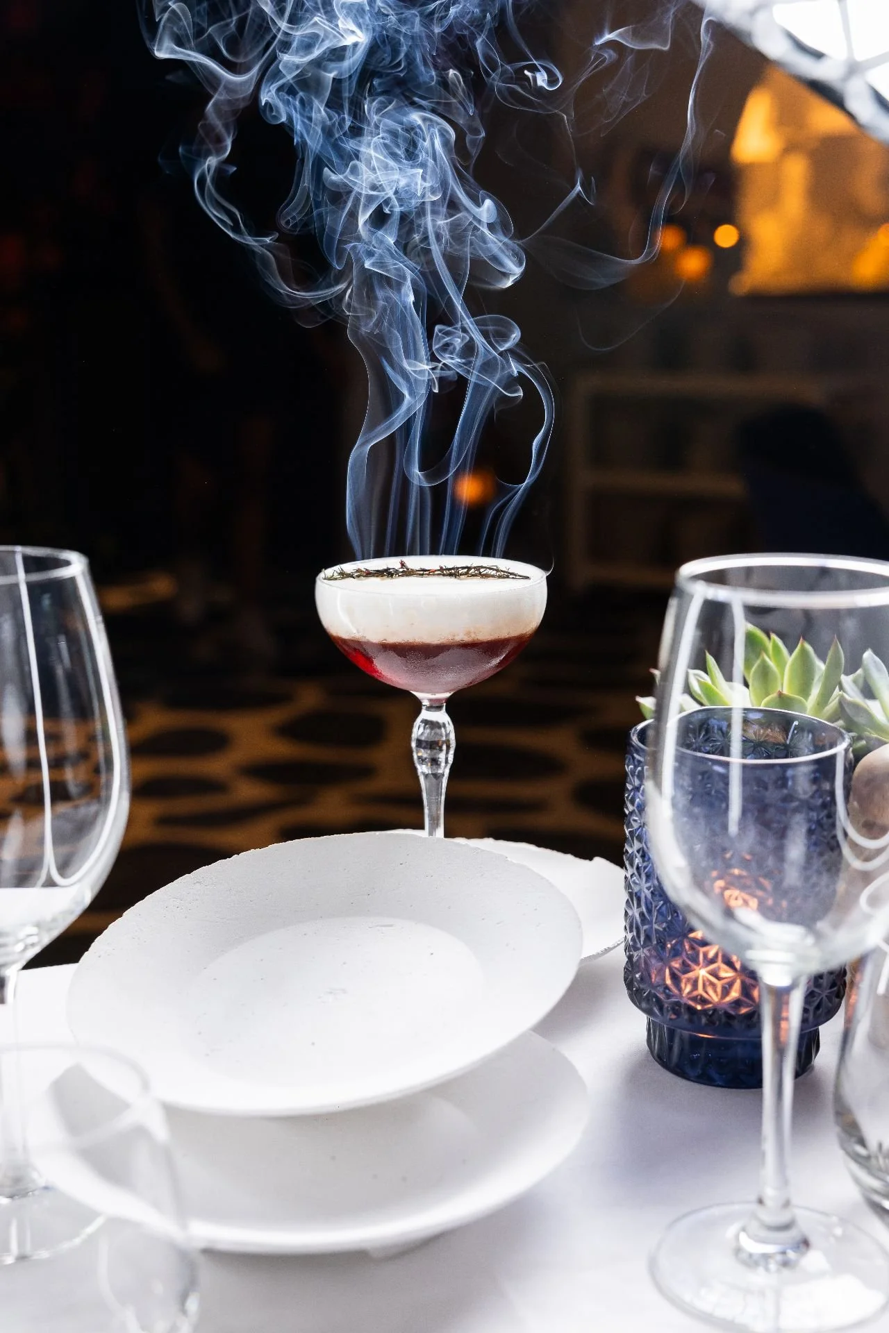A white, tiered cake stand with a hollow center, set on a table with empty champagne flutes and a glass of layered cocktail with smoke rising from it, and a small potted succulent plant.