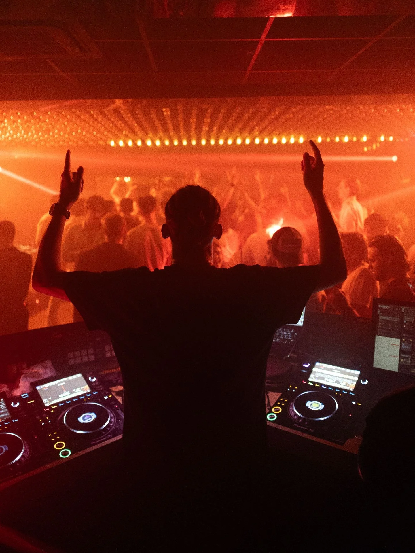 DJ performing at a nightclub with a crowd dancing in a red-lit environment and a DJ booth with turntables and equipment.