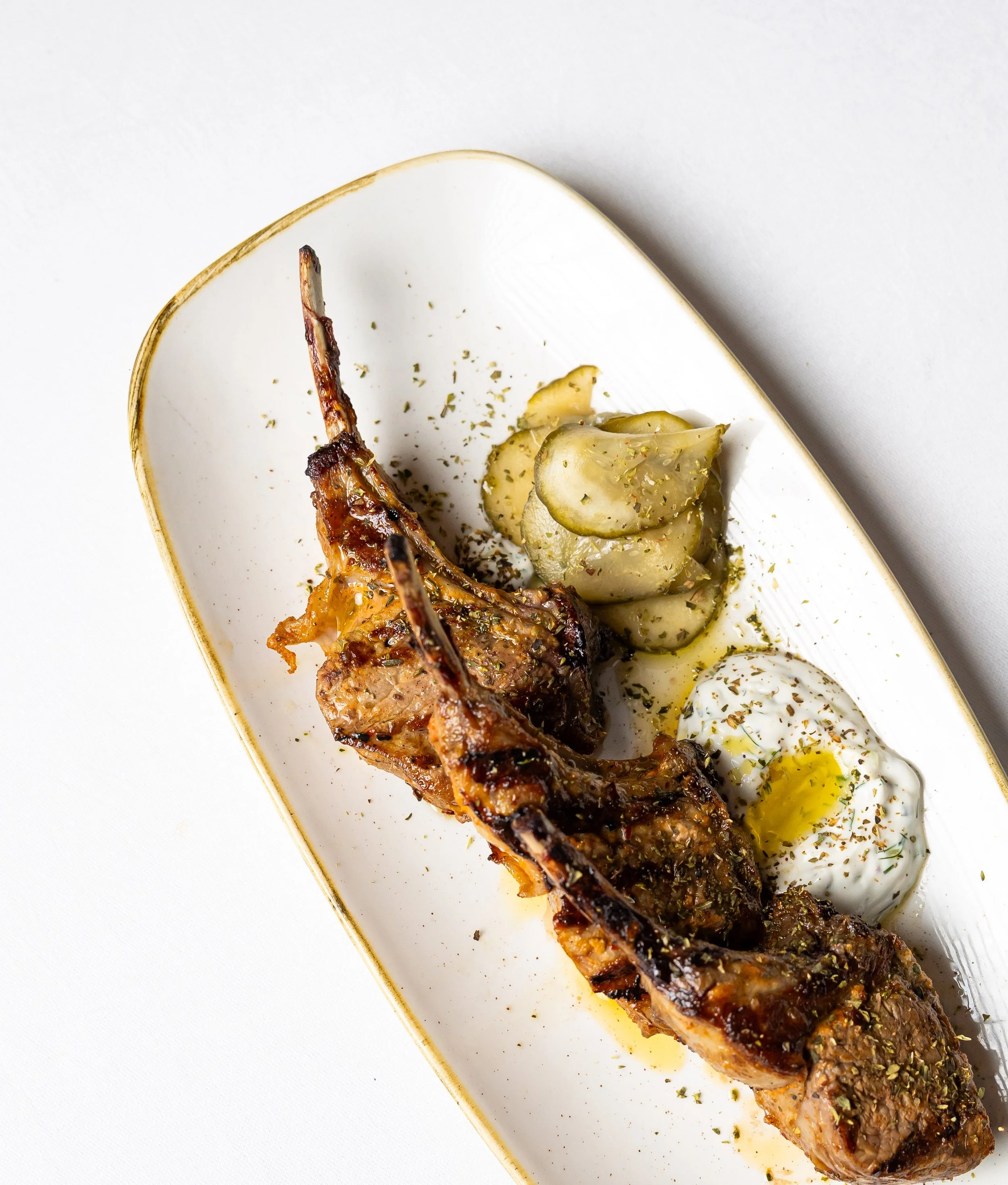 Close-up of a grilled meat skewer with herbs, served with sliced pickles, a boiled egg, and olive oil on a white plate with a gold edge, against a white background.