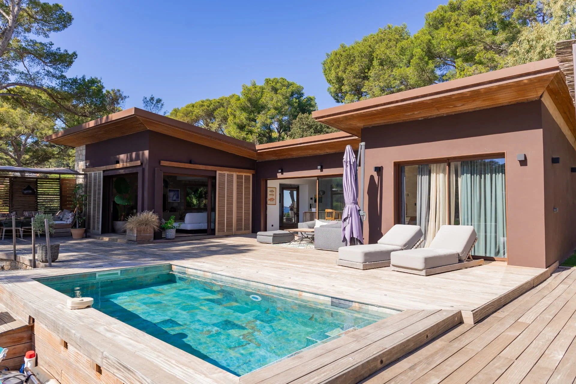 Modern house with outdoor pool, lounge chairs, and patio furniture surrounded by trees under a clear blue sky.