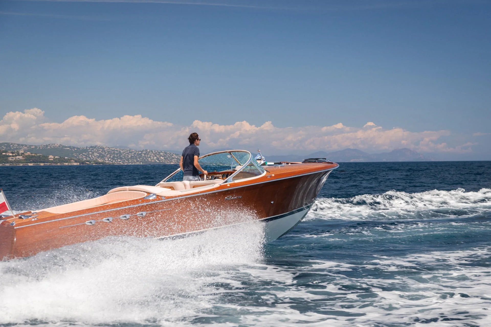 A man driving a wooden speedboat on the water with a scenic land and mountain background under a partly cloudy sky.