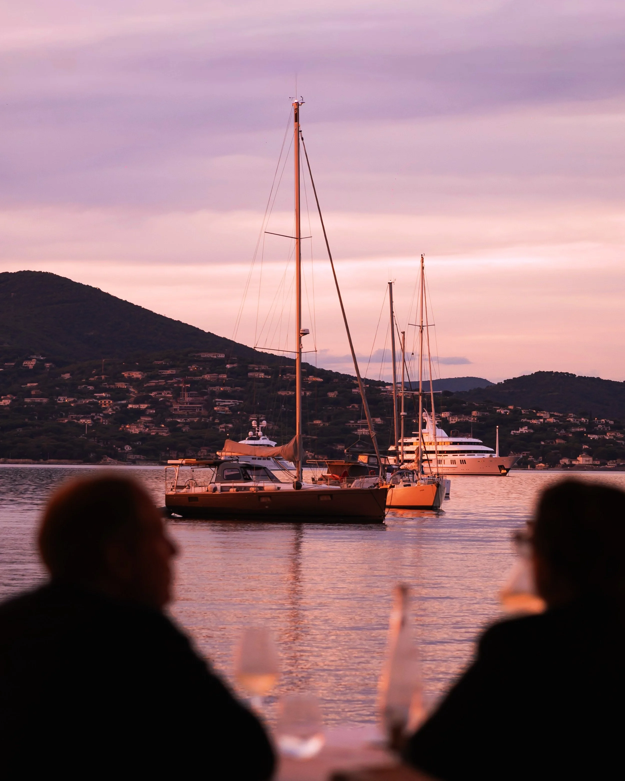 View of sailboats and a yacht moored in a harbor during sunset with reflective water, silhouetted figures, and a mountain backdrop.