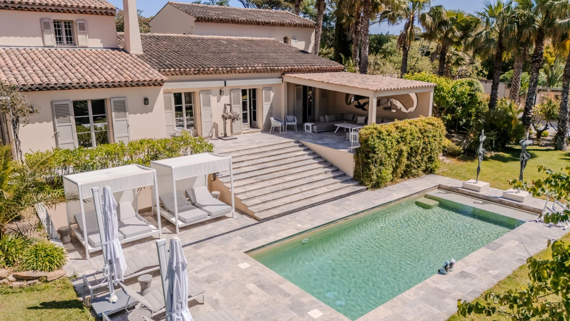 A backyard with a swimming pool, surrounding lounge chairs with umbrellas, a patio with steps leading to a covered seating area, and a house with tiled roof and white window shutters, with lush green trees and tall palm trees nearby.