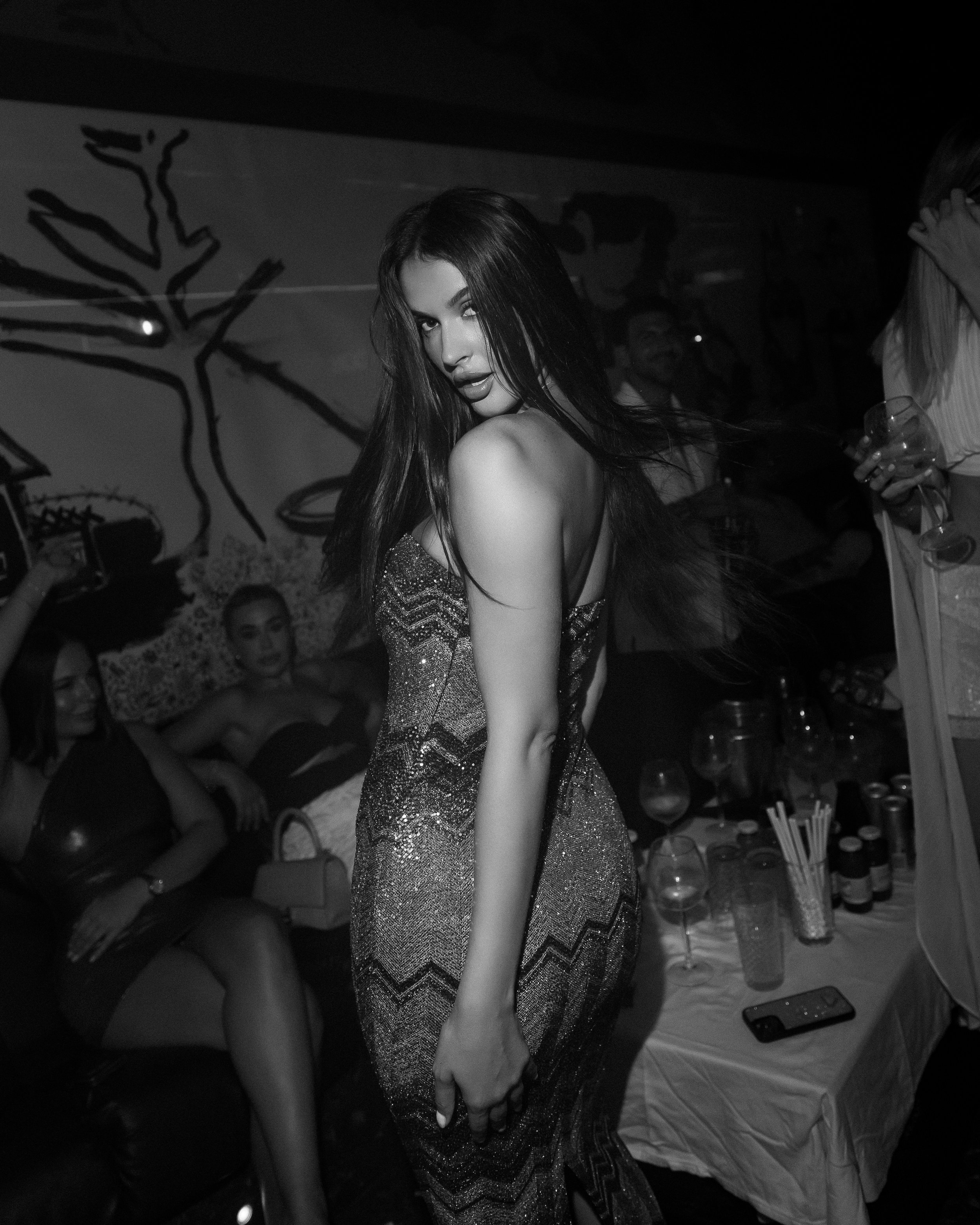 A woman with long hair looking over her shoulder, wearing a sparkly dress in a dimly lit room with people sitting on a black leather couch in the background.