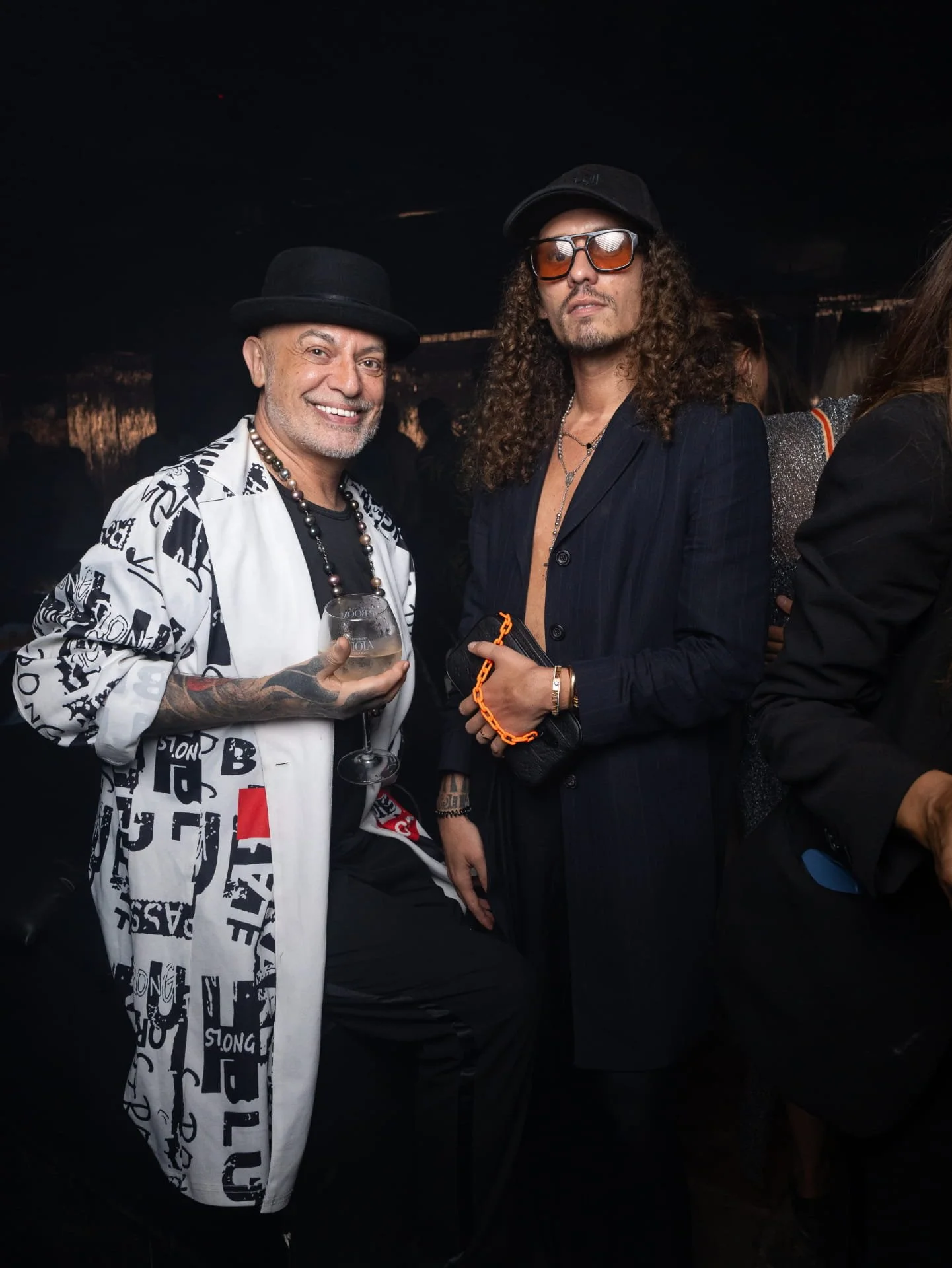 Two men at a social event. The man on the left is smiling, wearing a black hat, white shirt with black print, and holding a wine glass. The man on the right has curly hair, glasses, and is dressed in a black pinstripe suit with a necklace.