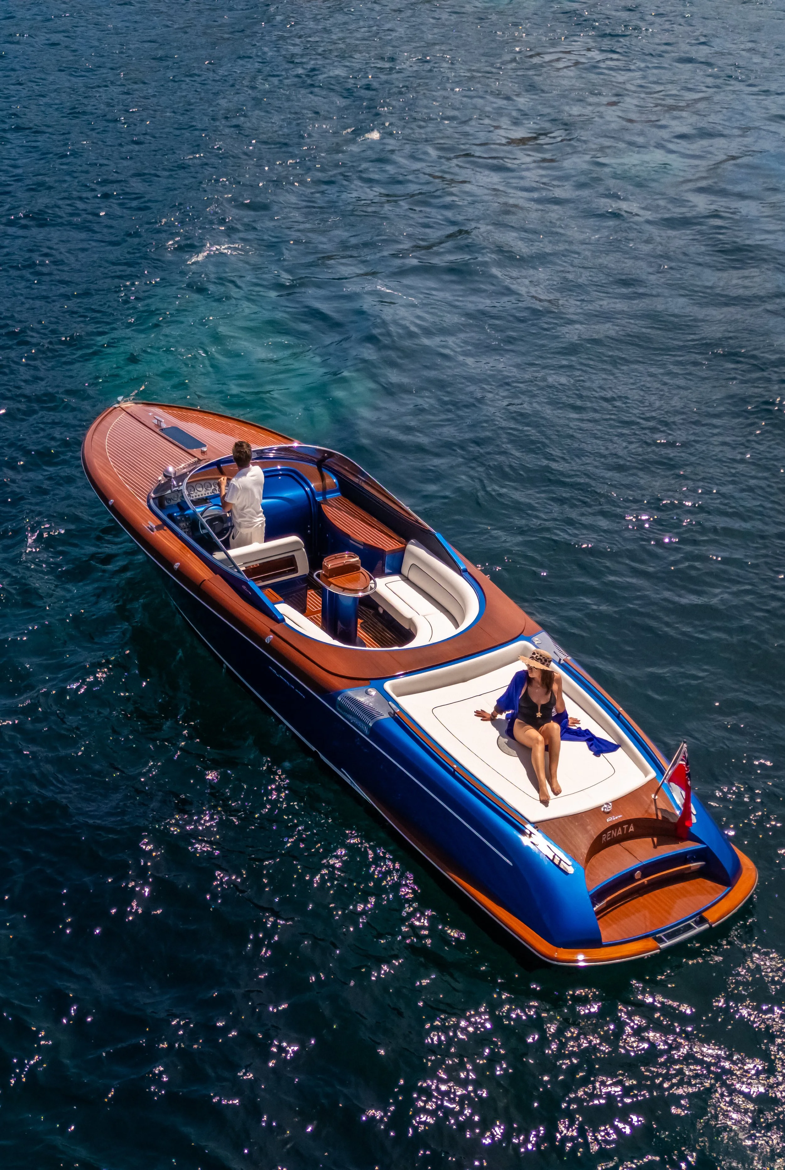 A luxury boat with a wooden deck and white seating area, floating on a calm blue ocean. A man is steering the boat, and a woman is sitting at the front, wearing a sunhat and enjoying the ride.