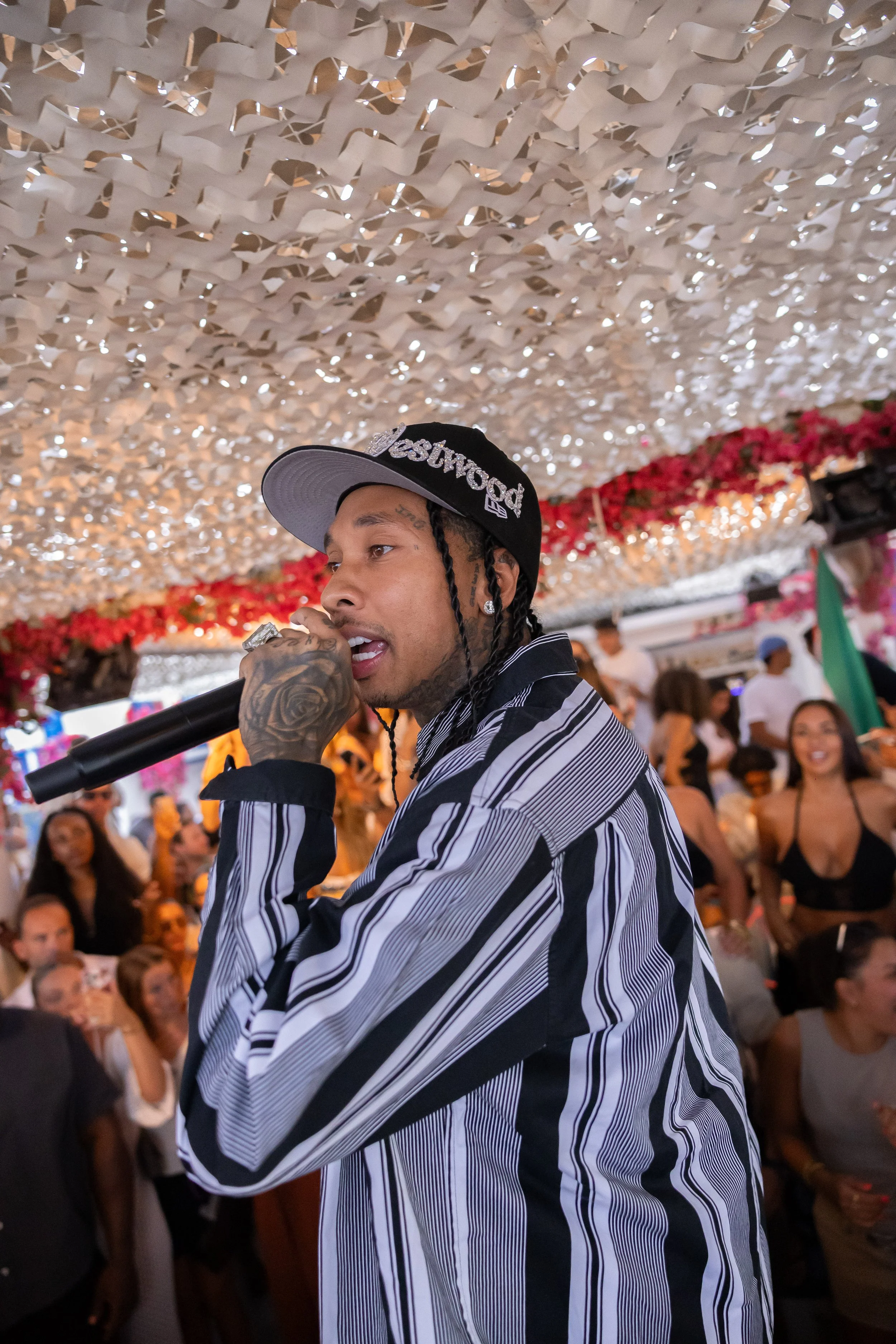A man with braided hair and tattoos, wearing a black and white striped shirt and a black cap with silver lettering, is holding a microphone at a crowded indoor event. The ceiling is decorated with white paper strips, and there are people in the backg