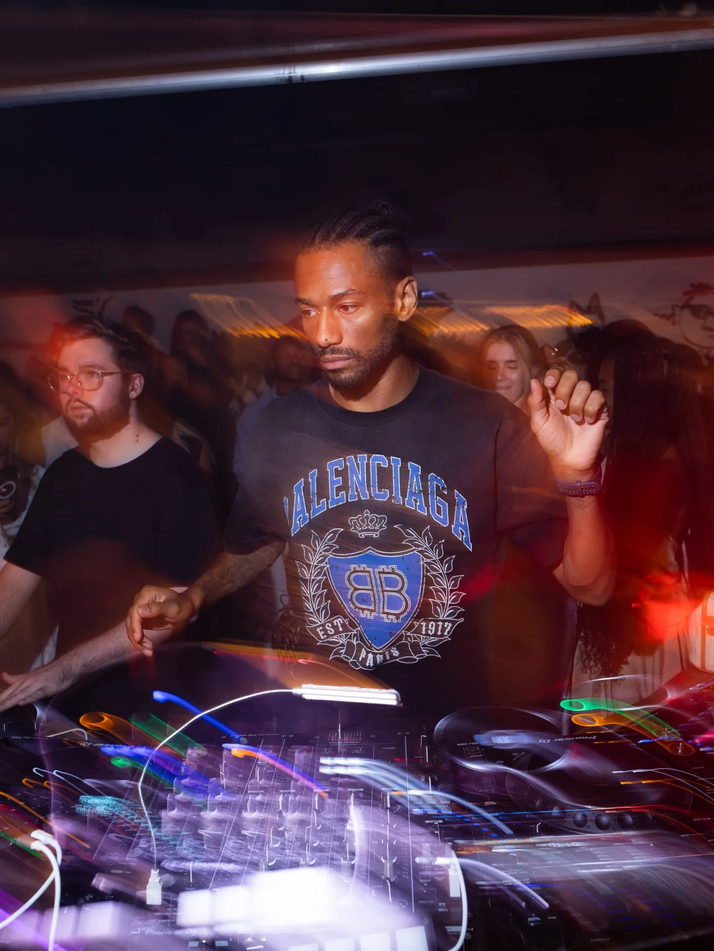 A man with braided hair and a beard wearing a black Balenciaga t-shirt is DJing at a party or nightclub with colorful lights and a crowd of people in the background.