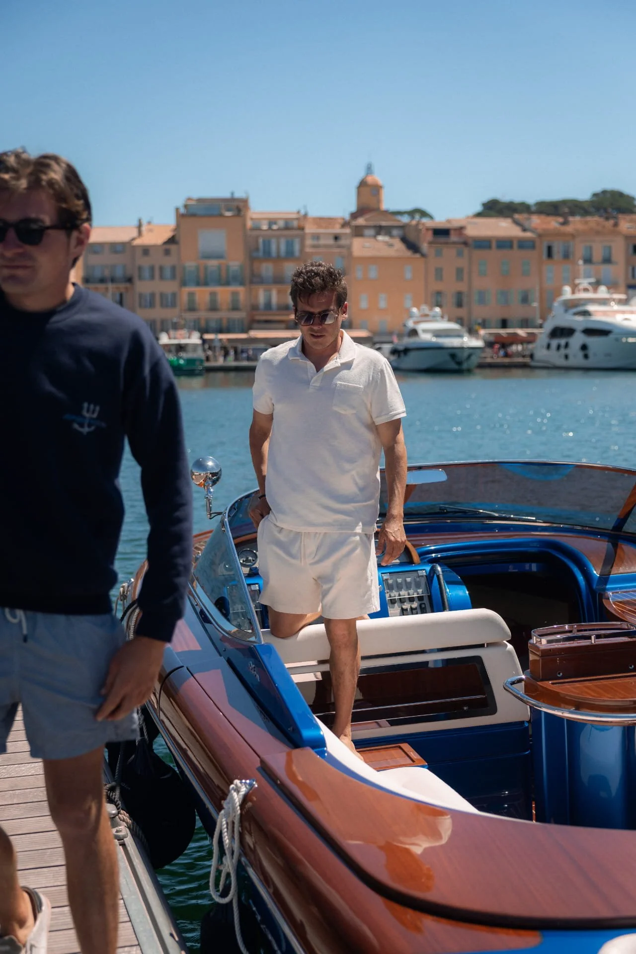 A man in white shorts and sunglasses standing on a yacht docked at a marina, with buildings and boats in the background.
