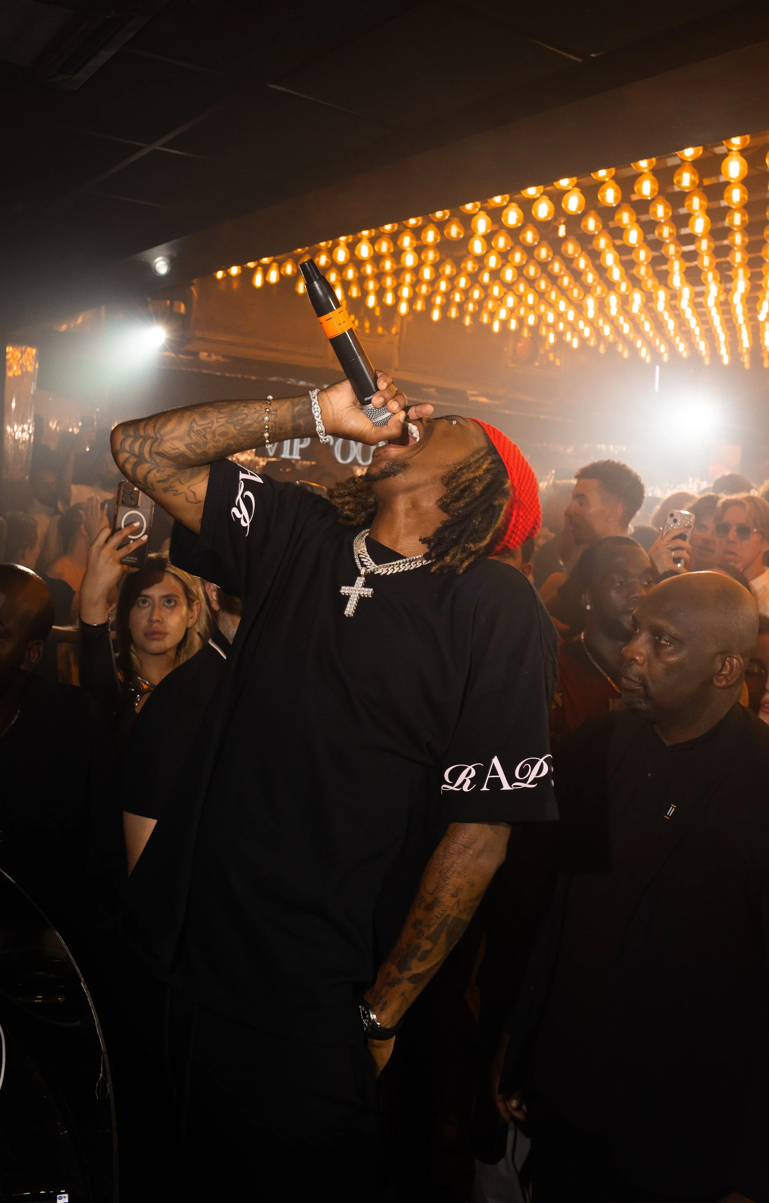 A man singing into a microphone at a crowded indoor event with bright orange ceiling lights.