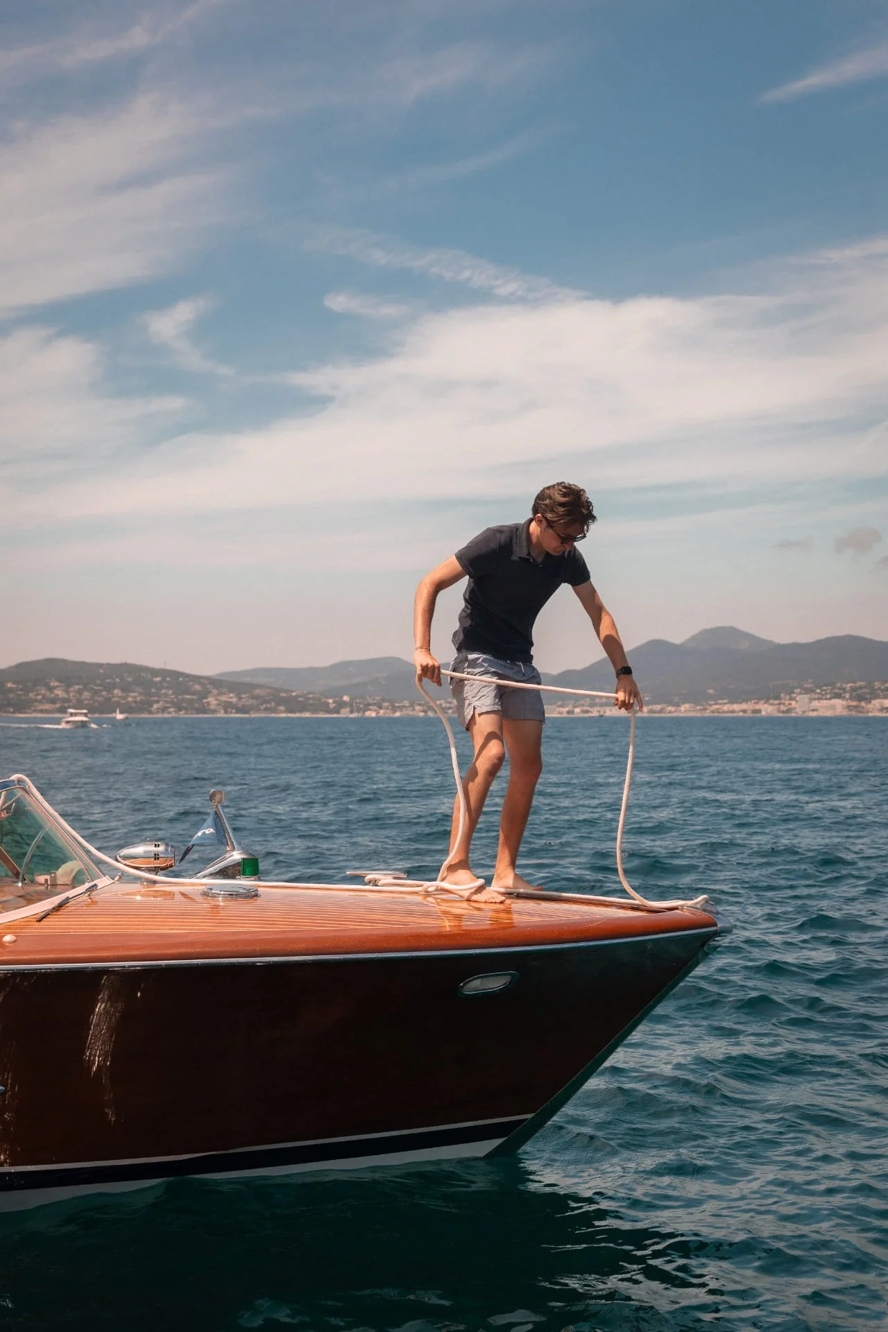 Young man in sunglasses and casual clothing on a yacht, handling a rope against the backdrop of water, distant mountains, and partly cloudy sky.