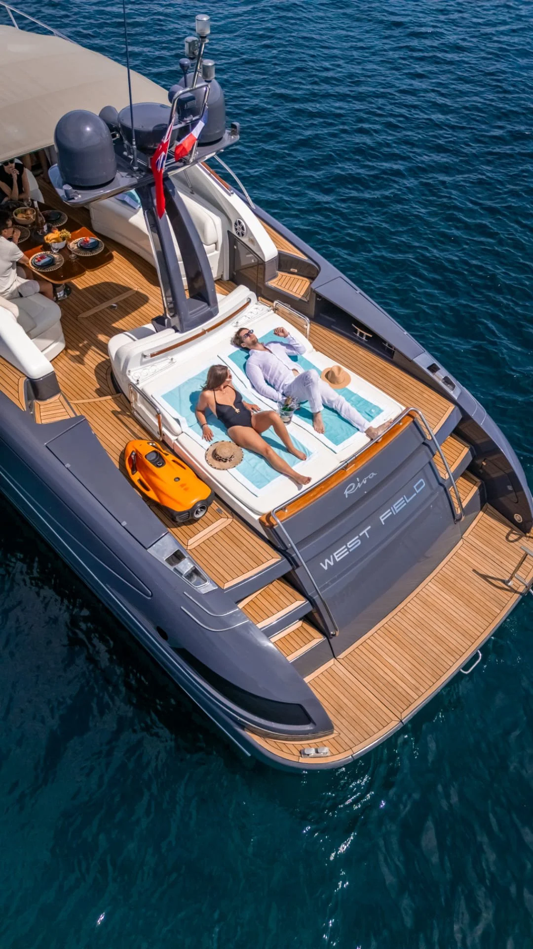 A luxury yacht with a wooden deck, two people lounging on a shaded area and sunbathing on a cushioned mat, surrounded by blue water.