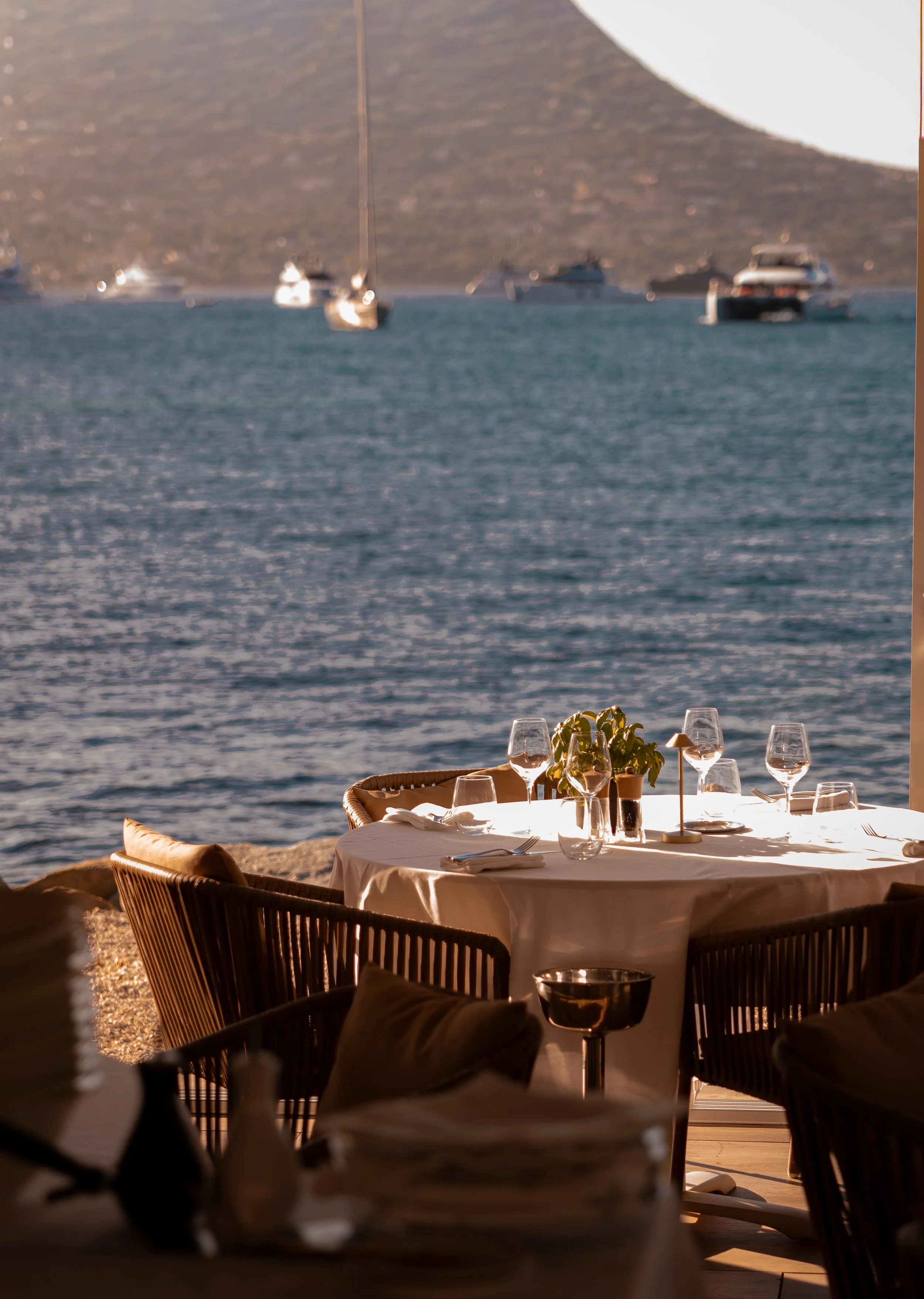 An outdoor restaurant table set with wine glasses, plates, and silverware overlooking a body of water with boats and a mountain in the background, during sunset or late afternoon.