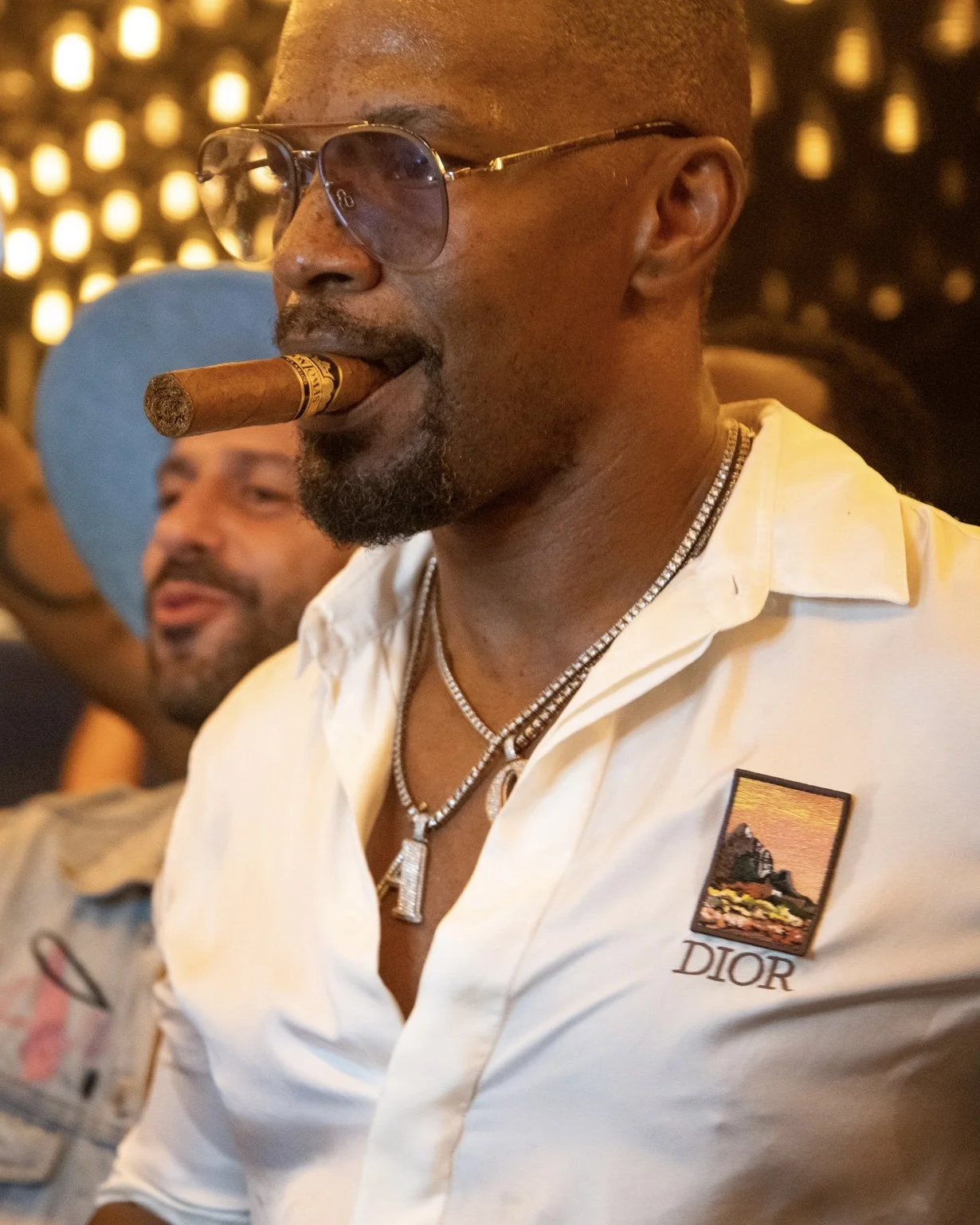 A man wearing sunglasses and a cigar in his mouth, dressed in a white shirt with a Dior logo and a patch, is seen at a social gathering. The background is decorated with warm, yellow lights.