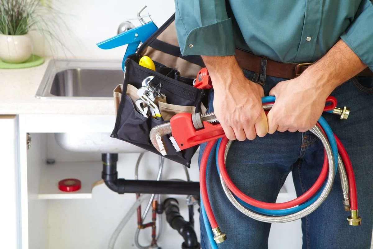 A person holding red and blue hoses with a pipe wrench, standing in a kitchen with a sink and a toolbox belt.