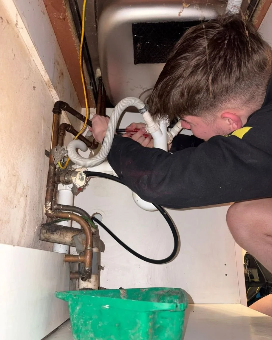 A person working on plumbing beneath a kitchen sink, with pipes, tools, and a green container nearby.