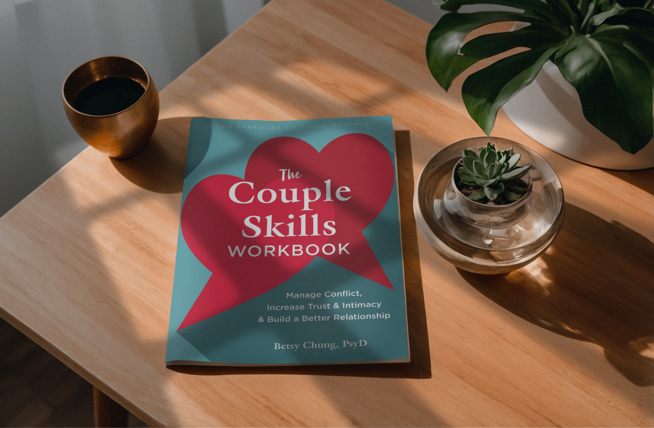 A self-help workbook titled 'The Couple Skills Workbook' is placed on a light wooden table. Next to it is a small potted succulent plant in a clear glass container. A large leafy green houseplant in a white pot is partially visible at the top right corner, and to the left of the workbook, there is a round, brown cup filled with black coffee. The sunlight is casting shadows on the table.