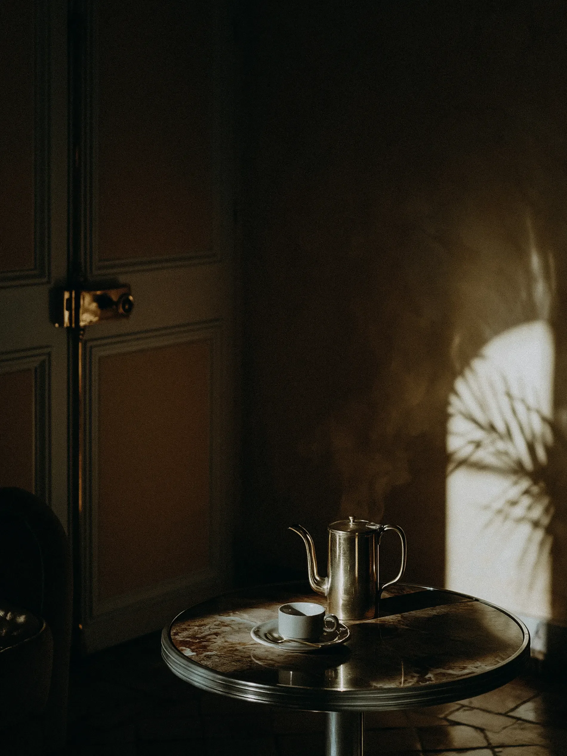A marble-topped round table with a coffee cup, saucer, and spoon, a metal pitcher, and a fire in the background casting shadows on a wooden-paneled wall.