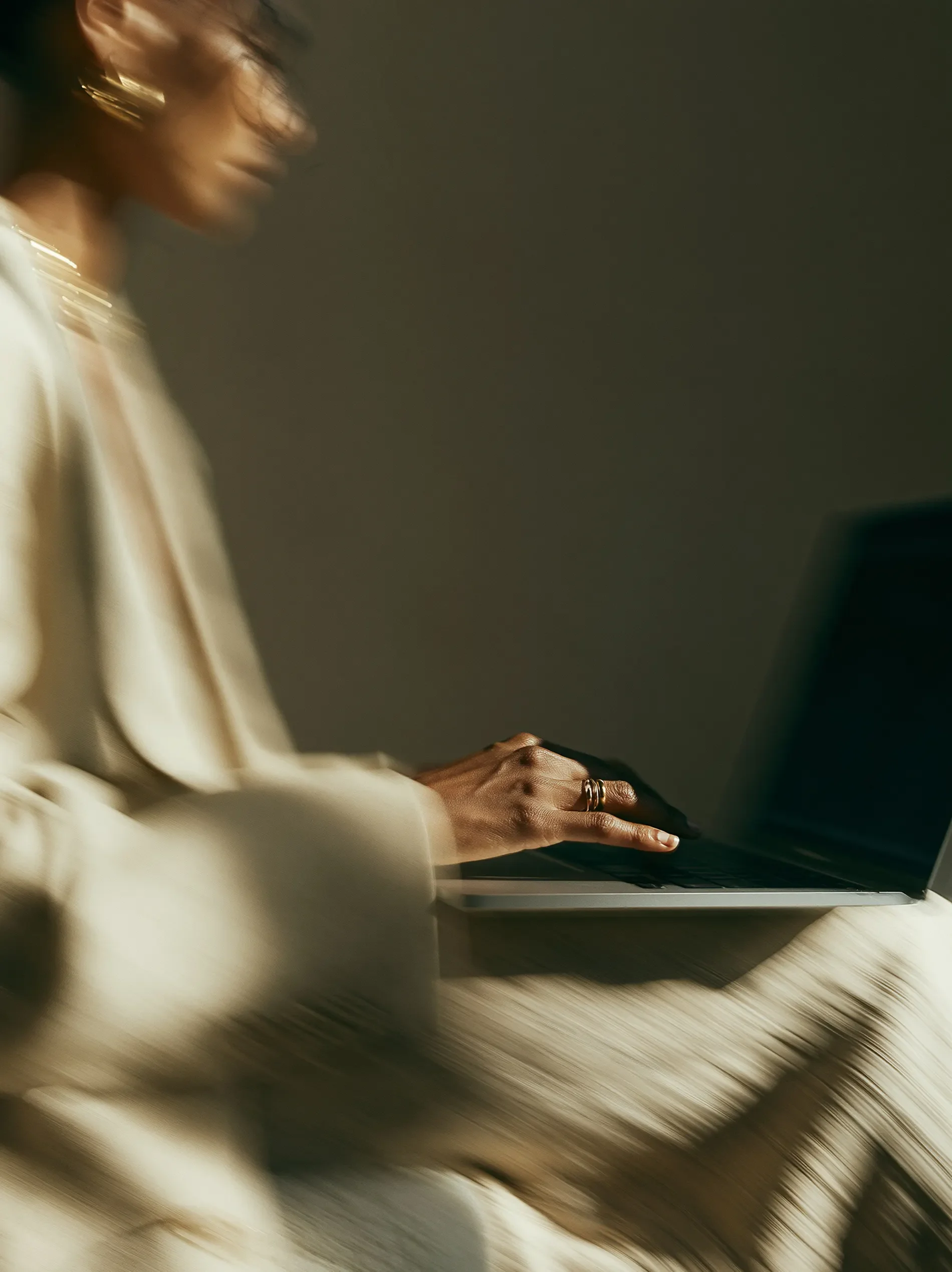 A person with dark skin wearing gold jewelry using a laptop, wearing a cream-colored long-sleeve top, with motion blur suggesting movement.