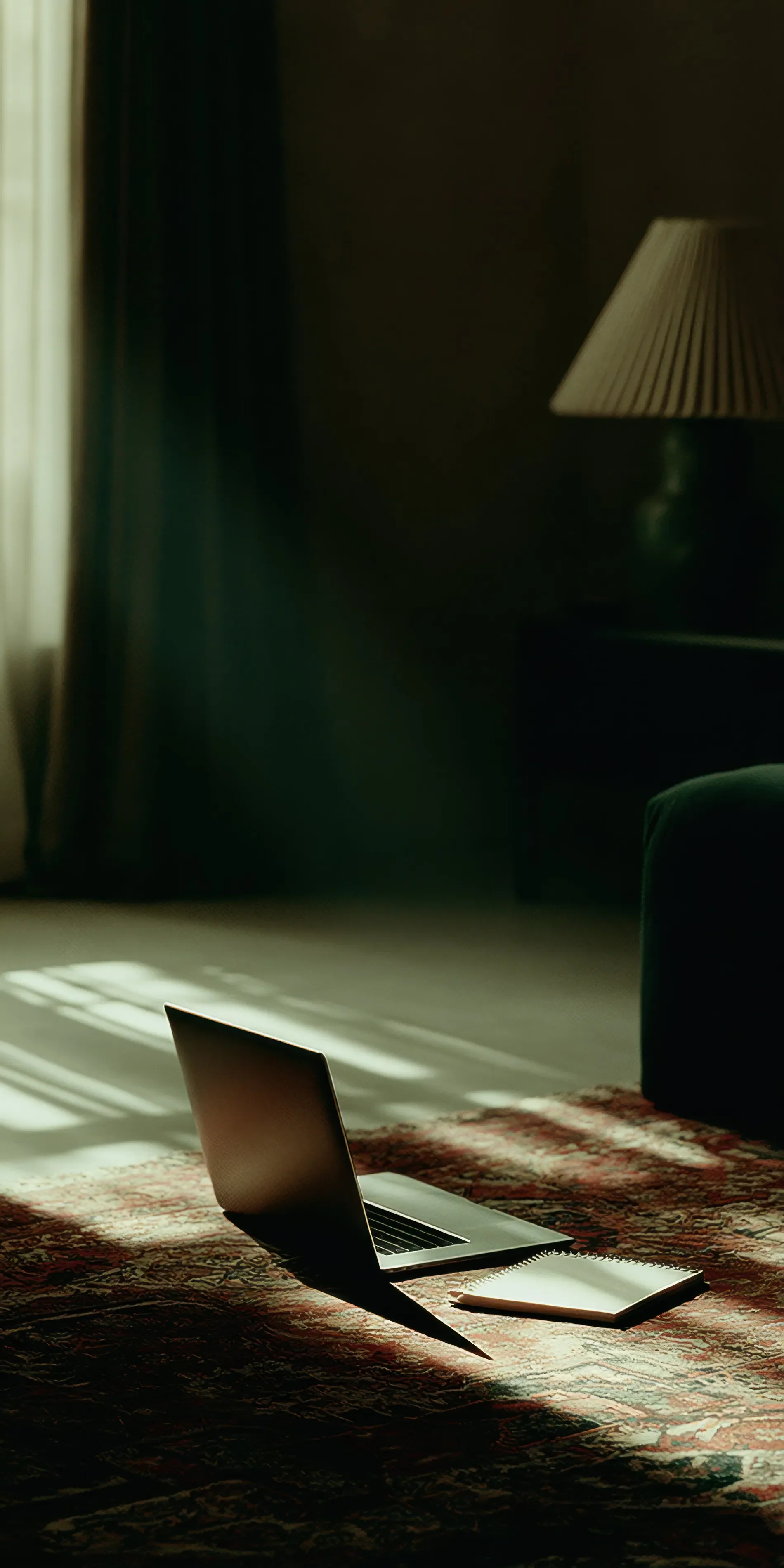 A laptop, a notebook, and a pen placed on a patterned rug in a dimly lit room with sunlight streaming through a window, casting shadows on the floor.