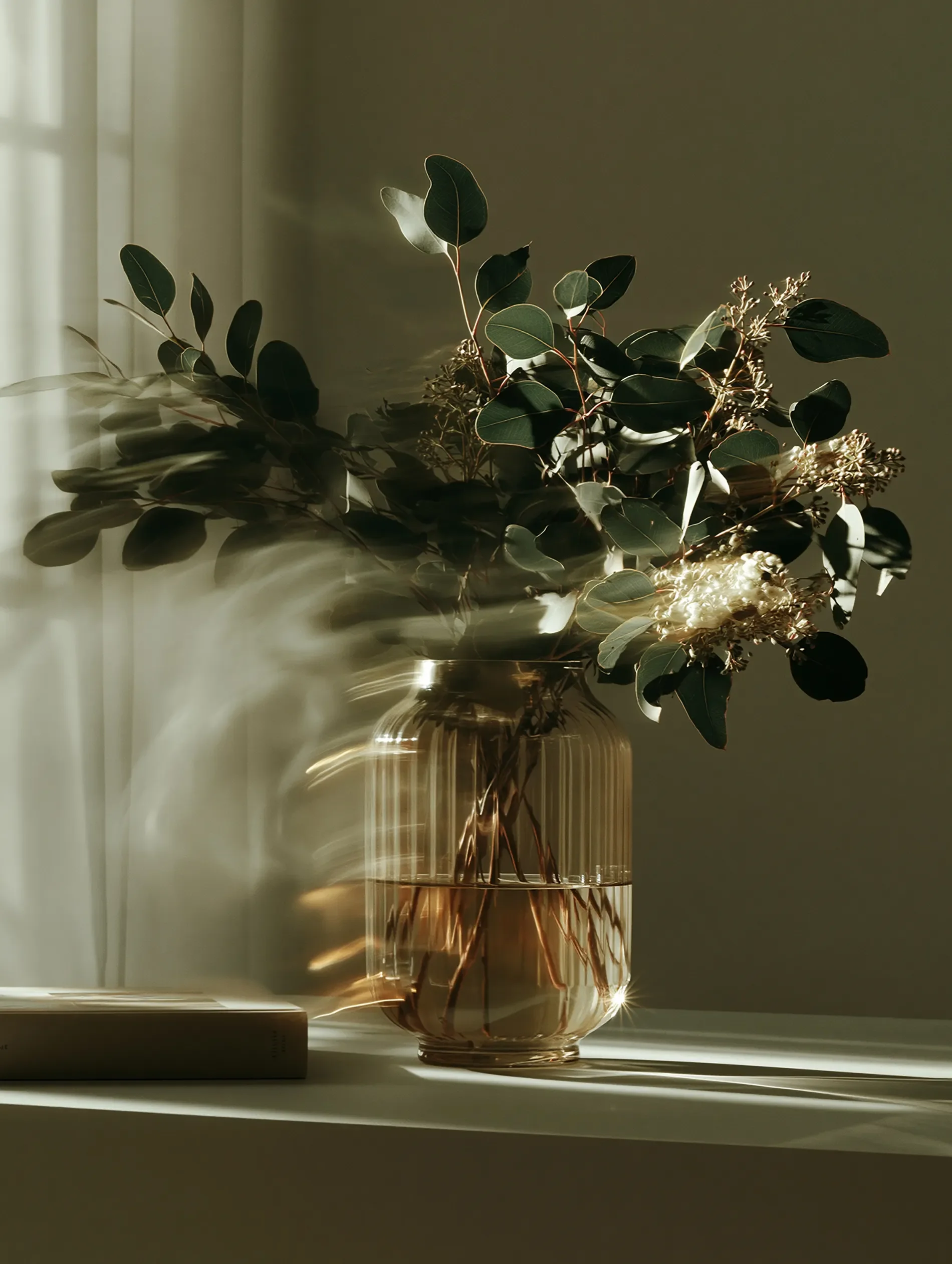 A glass vase with a bouquet of green leaves and small white flowers, illuminated by sunlight casting shadows on a white surface and wall.