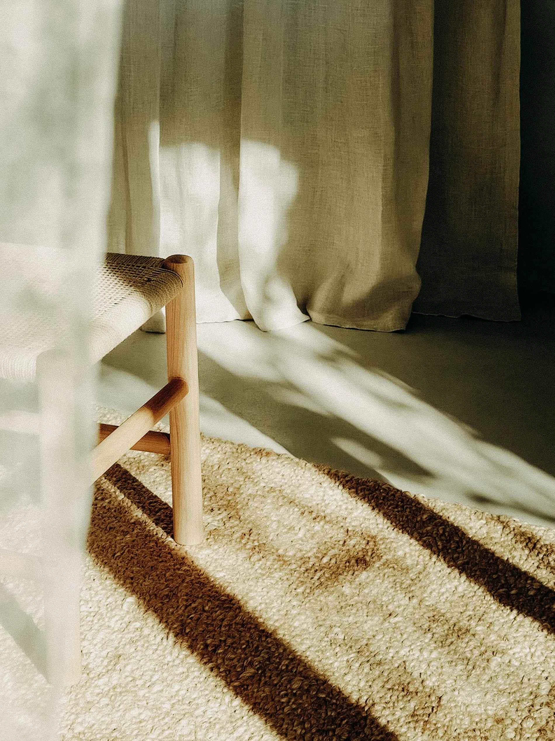 Close-up of light-colored and dark-colored striped rug on a white floor, with a wooden-framed woven chair beside curtains in the background.