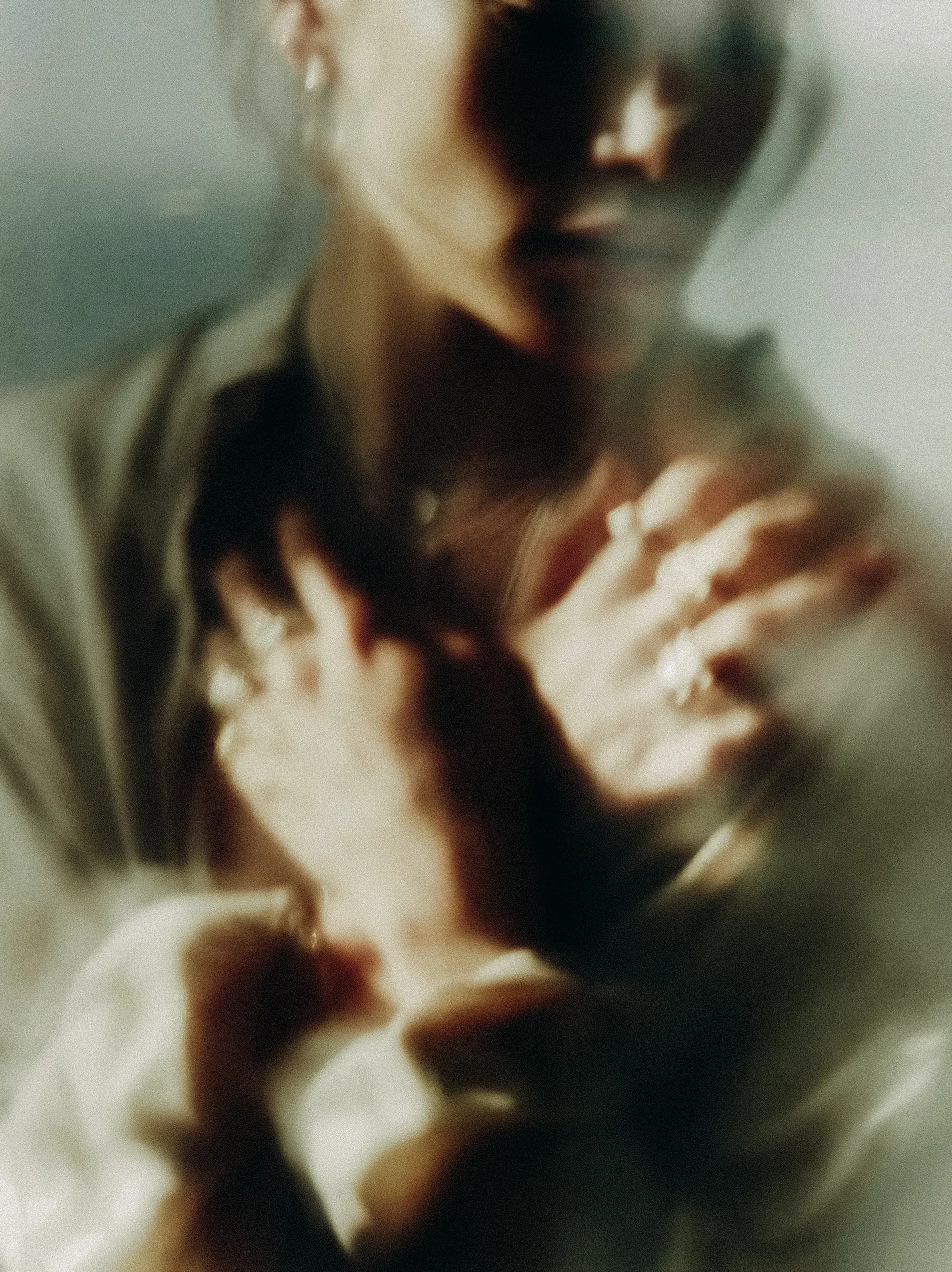 Close-up of a person's face with dark hair and dark makeup, with part of their hand visible, in a soft focus and muted colors.