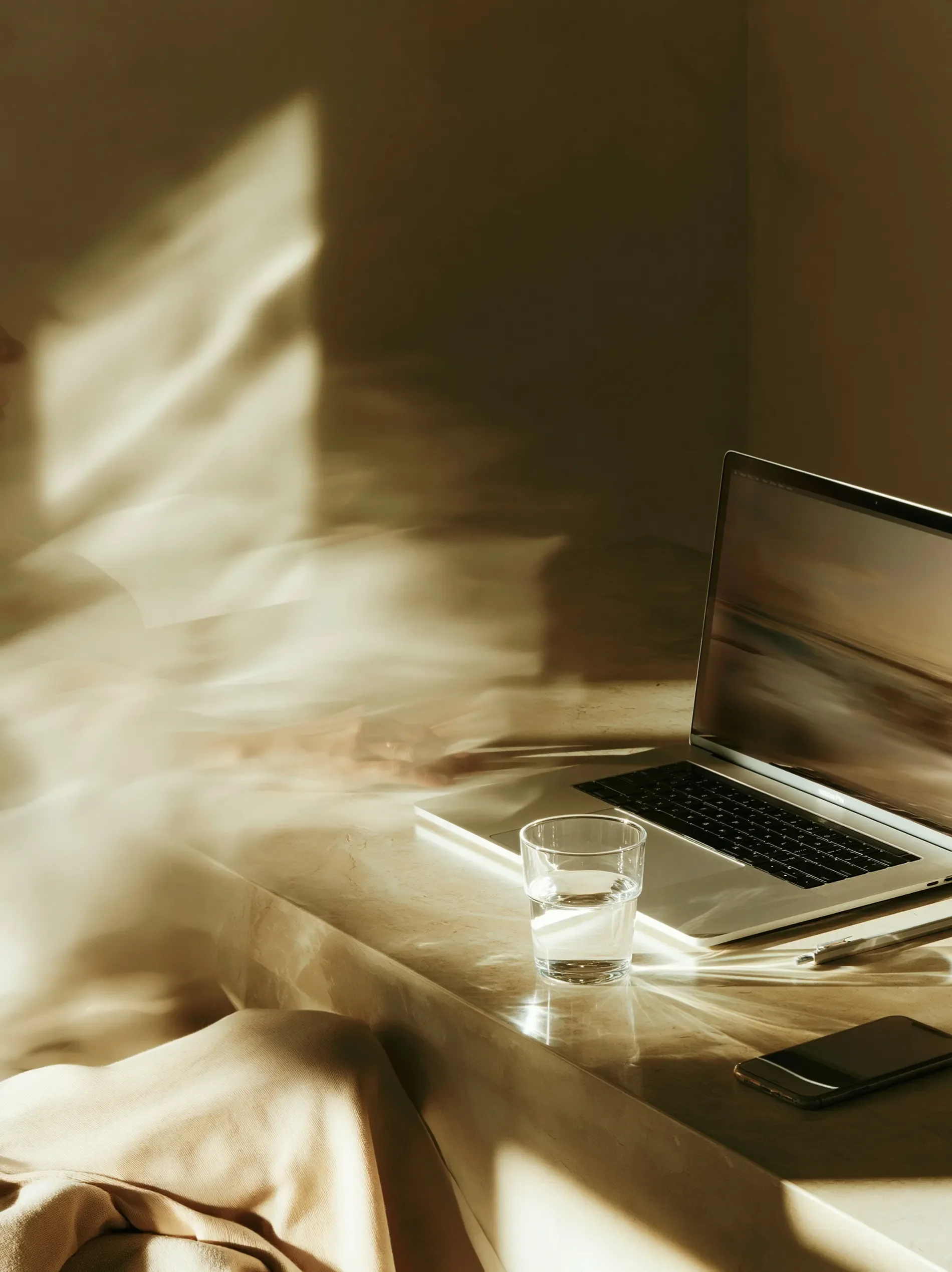 A laptop and a glass of water on a beige marble table, with sunlight creating bright patches and shadows on the surface. Part of a person's leg dressed in beige pants is visible in the bottom left corner.