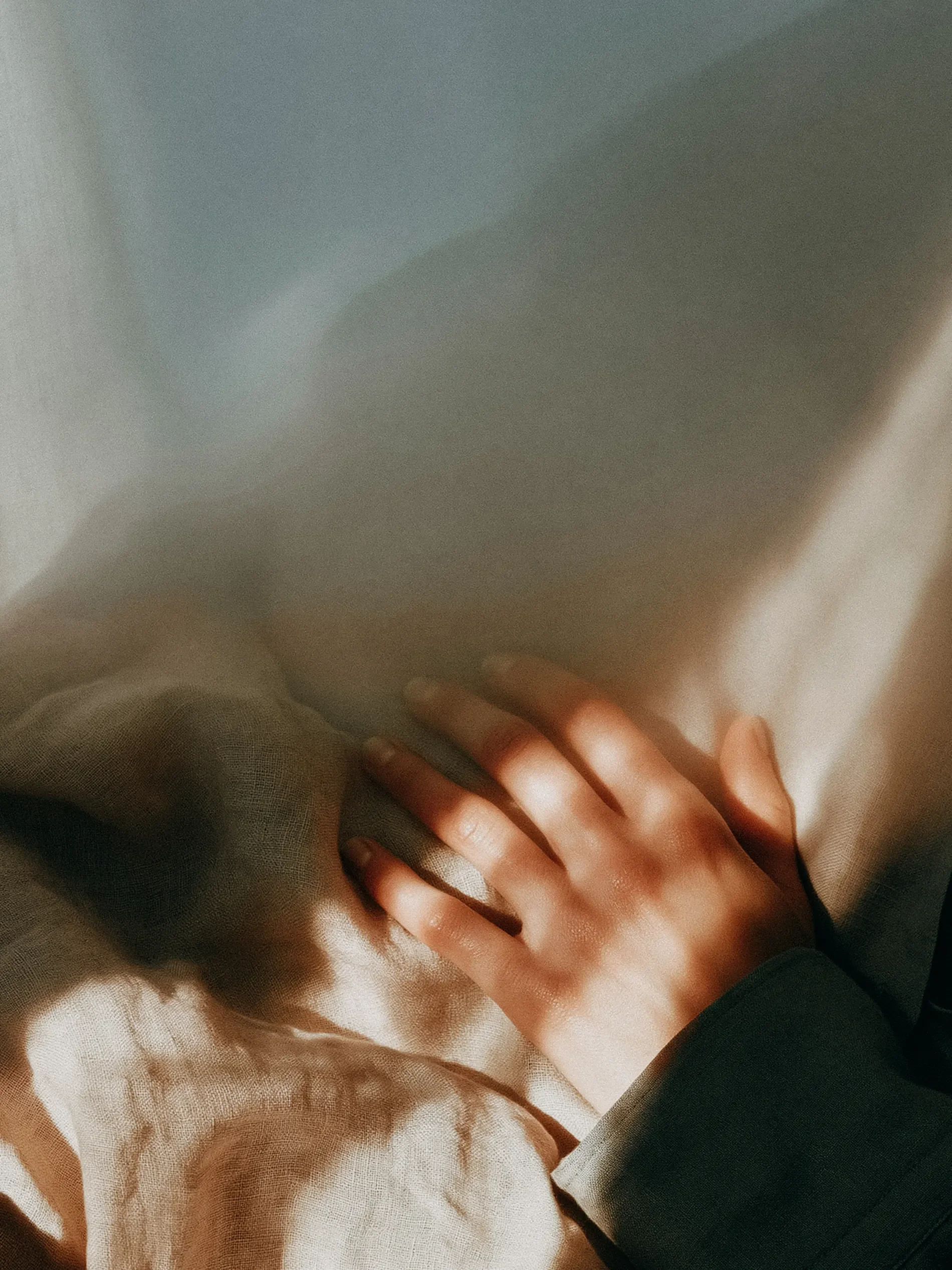 Close-up of a hand resting on a soft, beige fabric, possibly a blanket or cushion, with natural soft lighting.