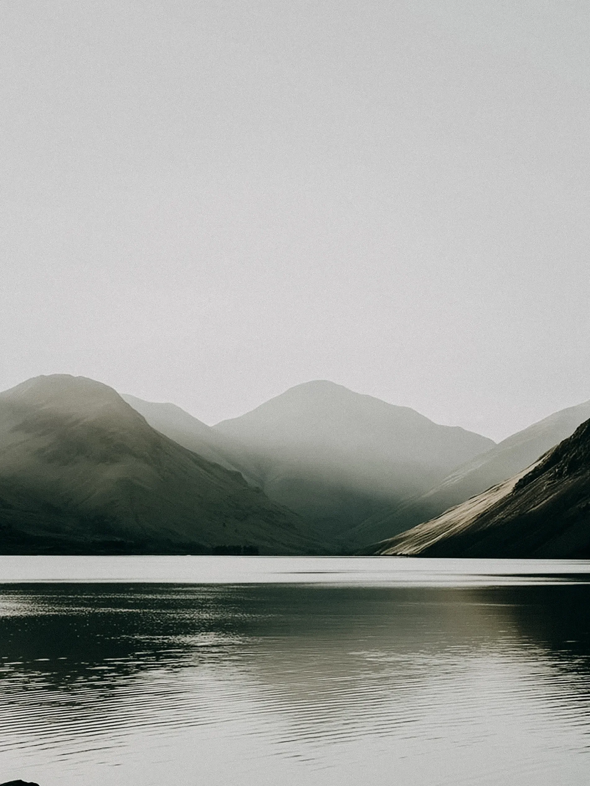 A calm mountain lake with dark water and gentle ripples, surrounded by mist-covered mountains in the background.