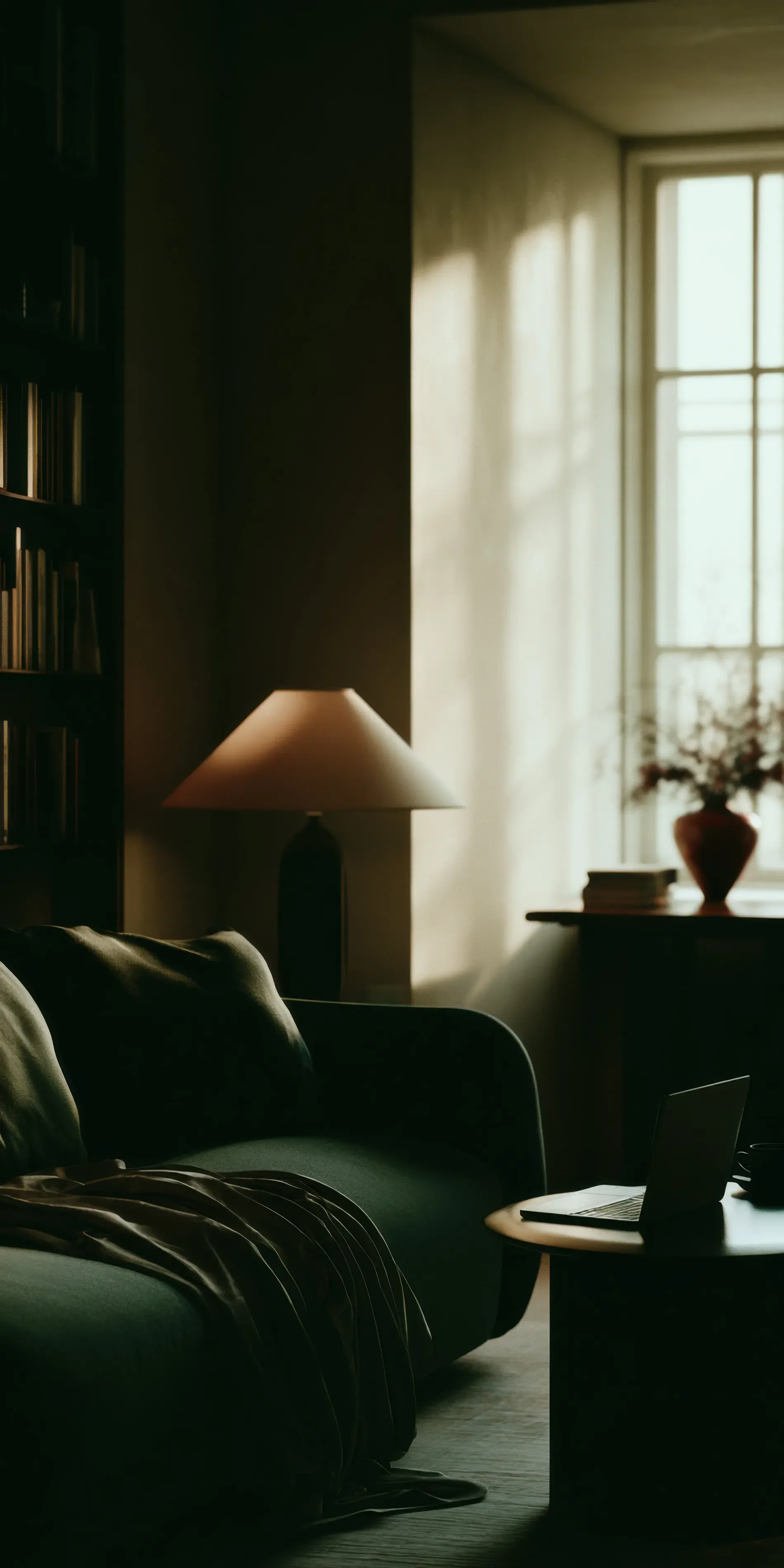 A cozy living room lit by sunlight from a large window, with a sofa, a side table with a laptop, a table lamp, a bookshelf, and a vase of flowers on another table.