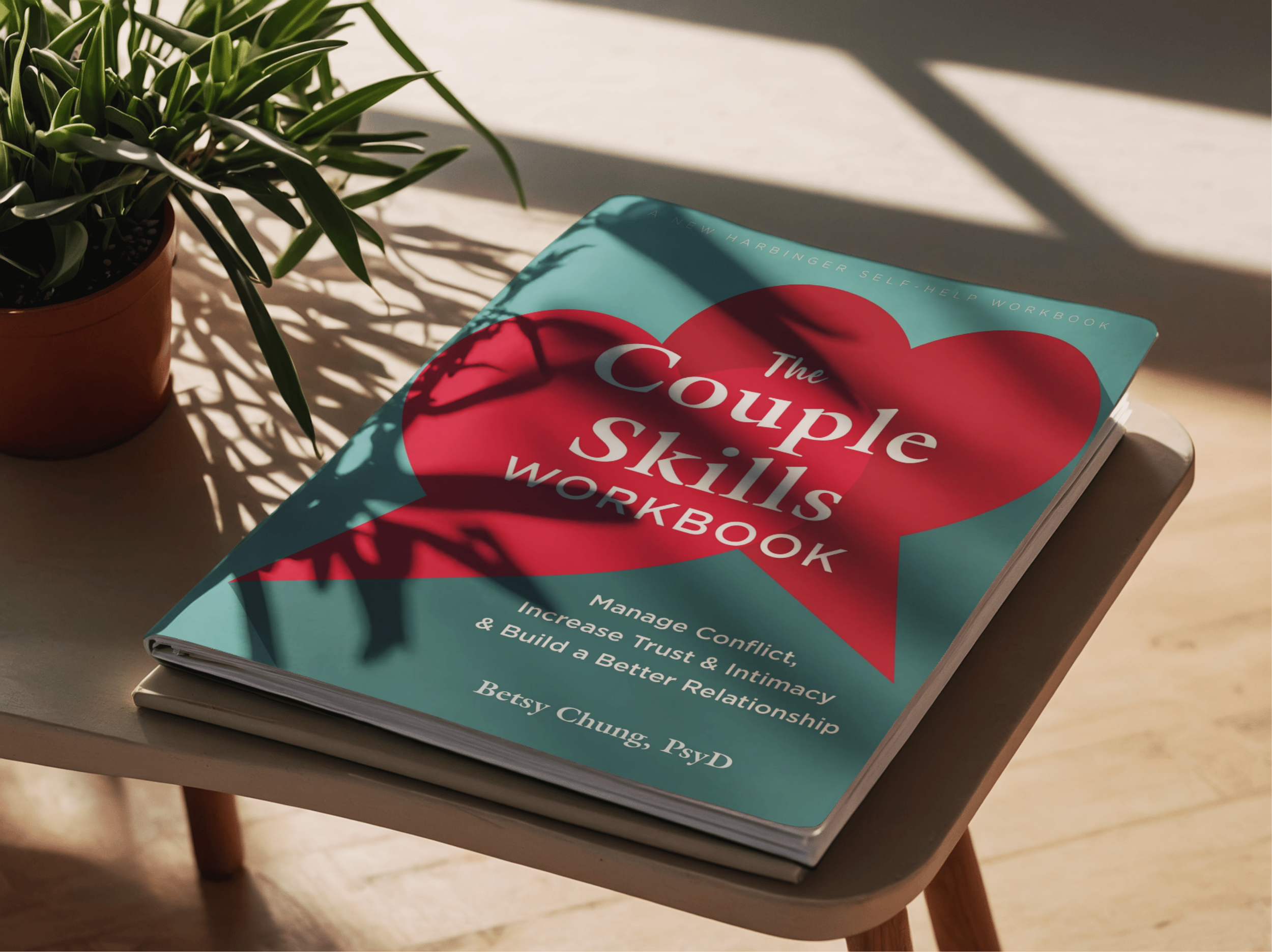 A book titled 'The Couple Skills Workbook' resting on a small table next to a potted plant. Sunlight creates shadows on the book and table.