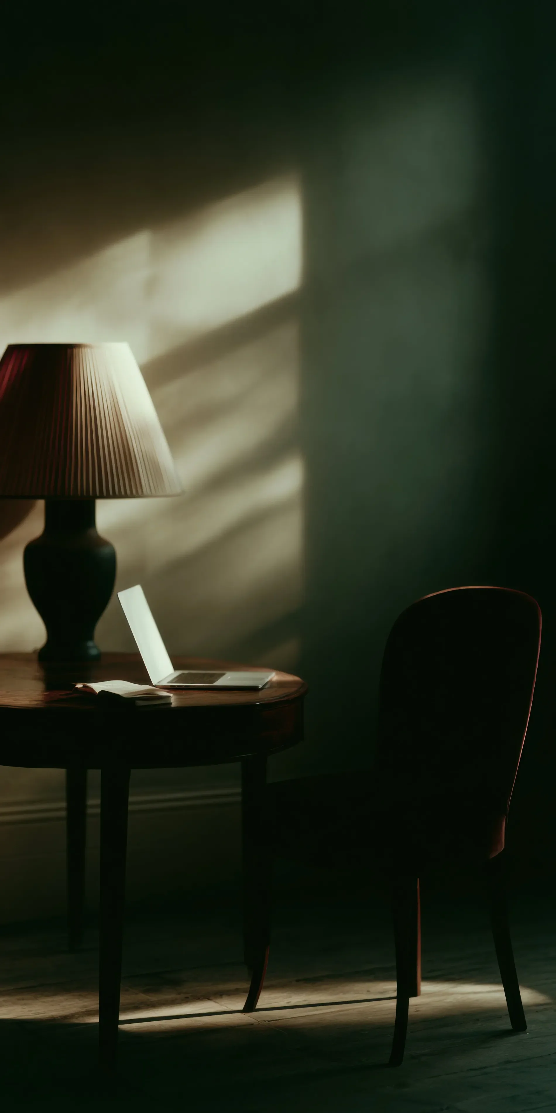 A dimly lit room with a wooden table holding a laptop, a notebook, and a pen. A classic table lamp with a striped shade provides warm light, while shadows cast patterns on the wall. A single dark chair is placed beside the table.