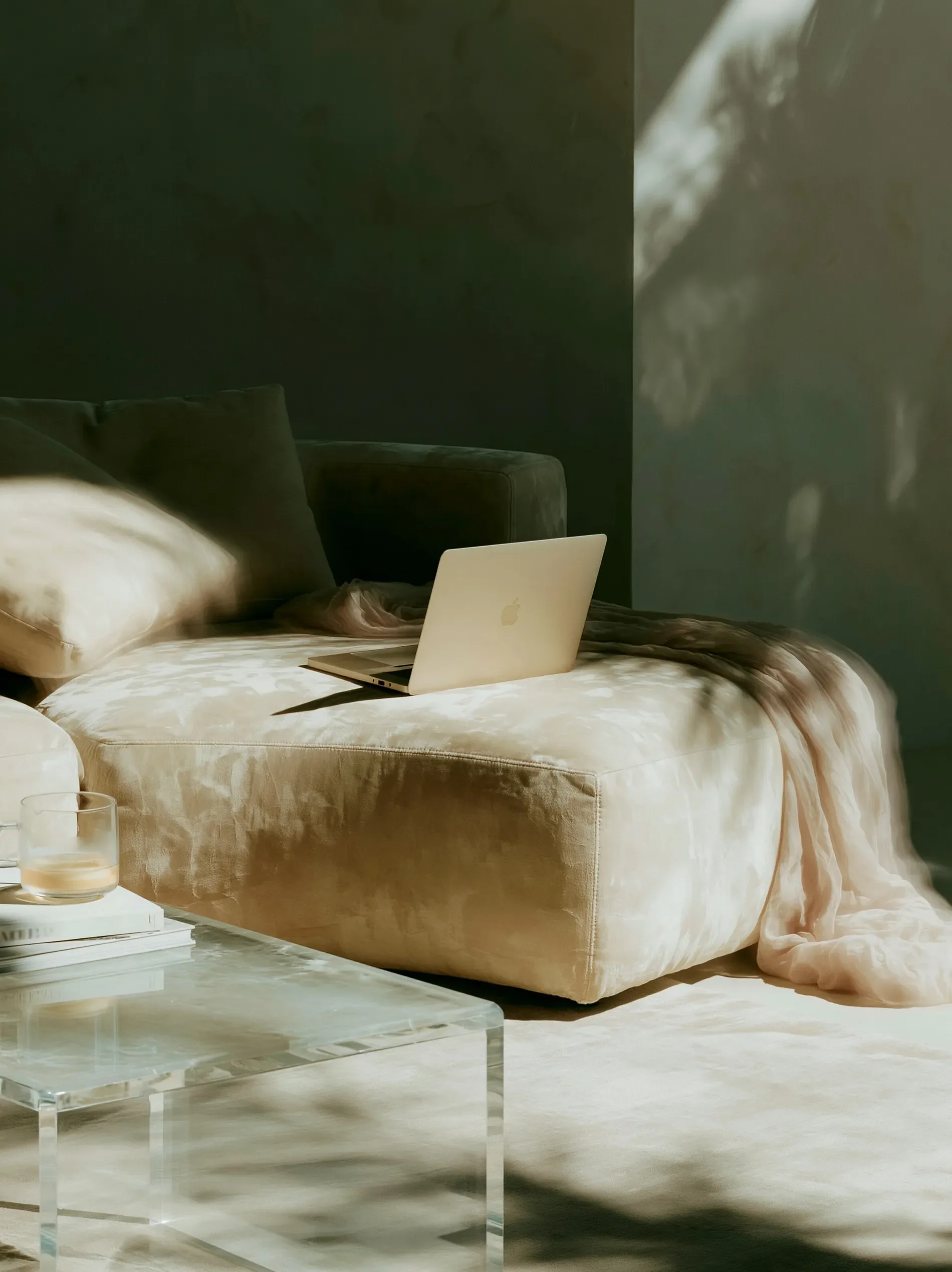 A cozy bedroom with a beige velvet bed, pillows, a gold throw blanket, a white MacBook on the bed, a glass of water, and a glass-topped coffee table, with a dark wall and soft natural light.