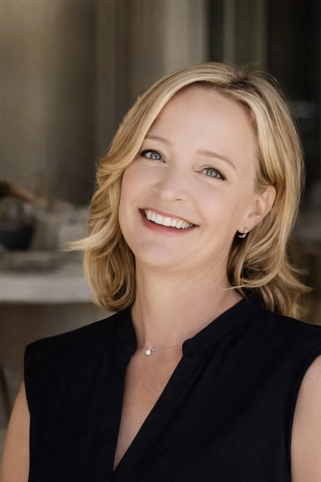 A woman with blonde hair smiling, wearing a black top and a necklace with a small pendant, in an indoor setting.
