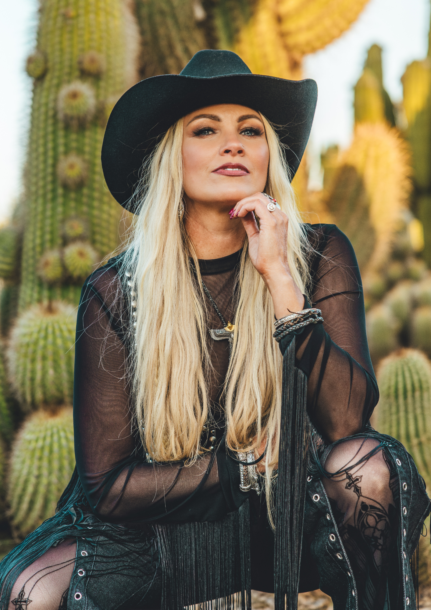 A woman with long blonde hair wearing a black hat and sheer black clothing, sitting on a cactus in a desert environment.