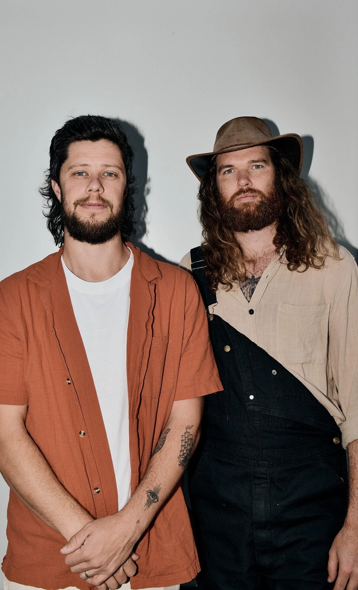 Two men stand together against a white background. The man on the left has dark, wavy hair, fair skin, a beard, and is wearing a reddish-brown short-sleeve button-up shirt over a white t-shirt. The man on the right has long curly hair, a beard, lighter skin, and is wearing a beige shirt with rolled-up sleeves, a brown hat, and black overalls.