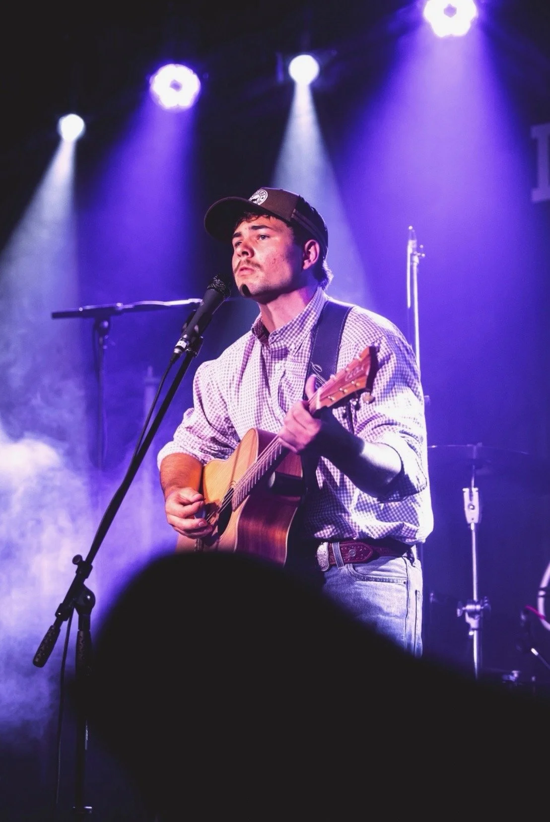 Musician performing on stage with guitar, wearing a baseball cap and plaid shirt, under purple stage lights, with microphone in front.