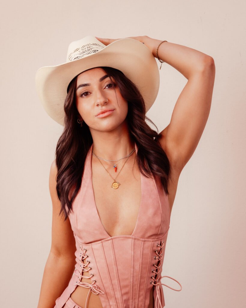 A young woman with long dark hair wearing a large beige sun hat, a pink corset-style top, and layered necklaces, posing with one arm raised and her hand on her hat against a neutral background.