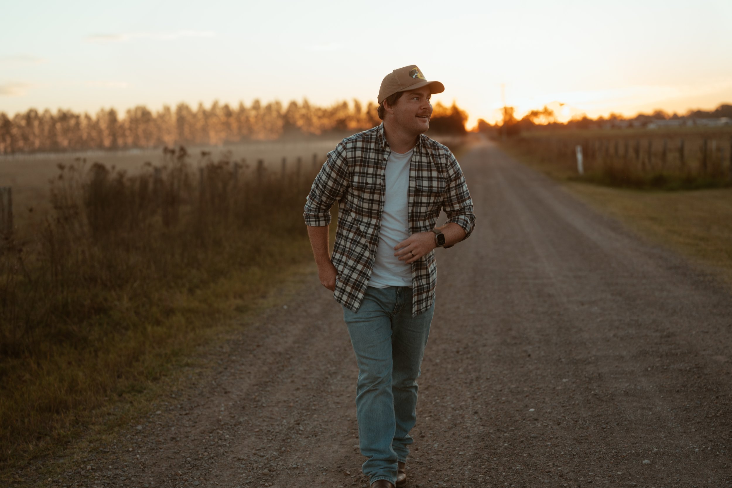 A man walking along a dirt road during sunset, wearing a plaid shirt, jeans, and a cap, with a watch on his wrist.