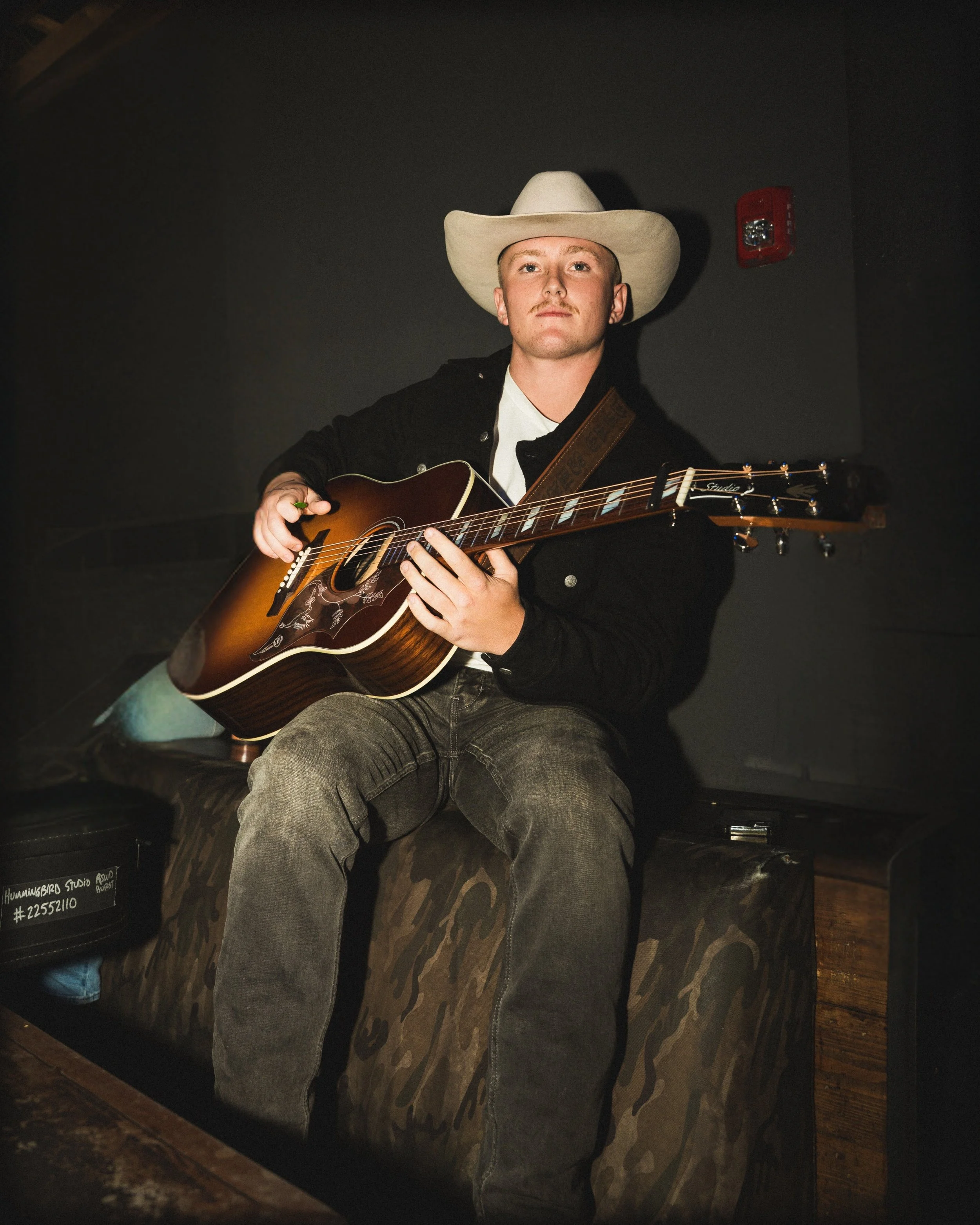 A man wearing a cowboy hat and a black jacket playing an acoustic guitar on stage, with dark background and stage lighting.
