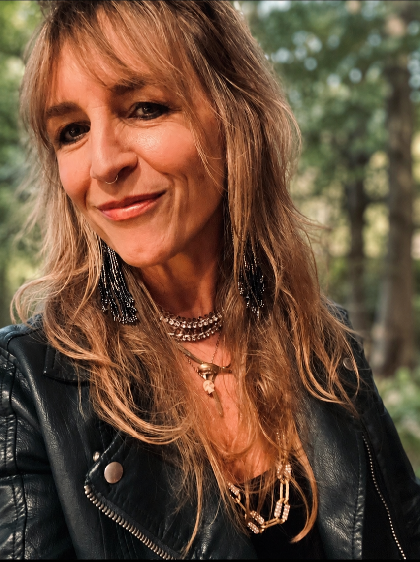 Close-up of a woman with wavy, reddish-brown hair wearing a black leather jacket and layered jewelry, outdoors with green trees in the background.